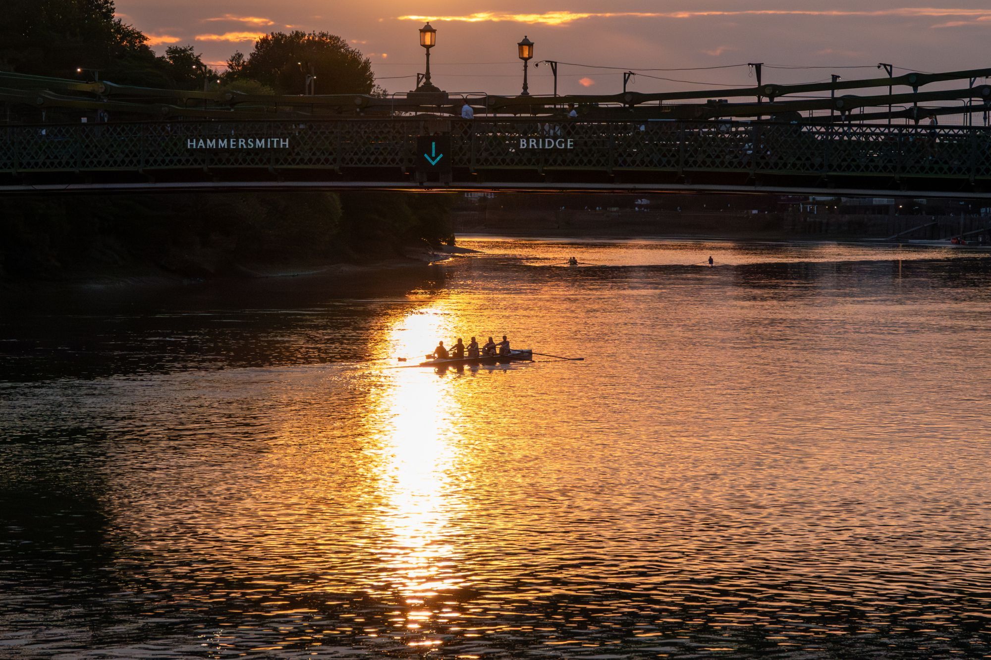 A sunset view under Hammersmith Bridge, there's an arrow pointing down on the bridge, directly beneath that is the sun reflected in a long line on the river, and people rowing a boat