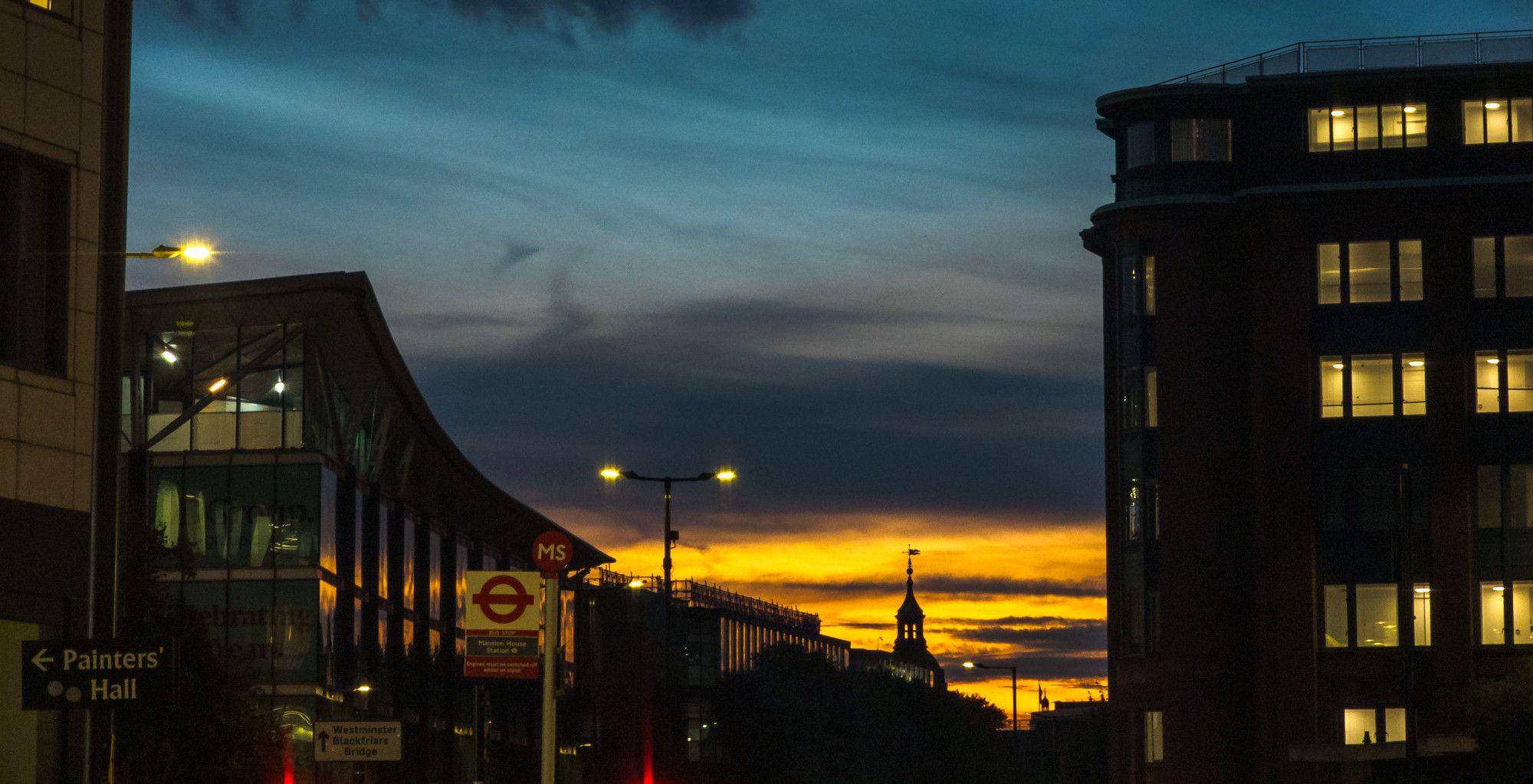 Sunset between city buildings, with a tower silhouetted against it. There's a sign on the right saying "Painters' Hall"