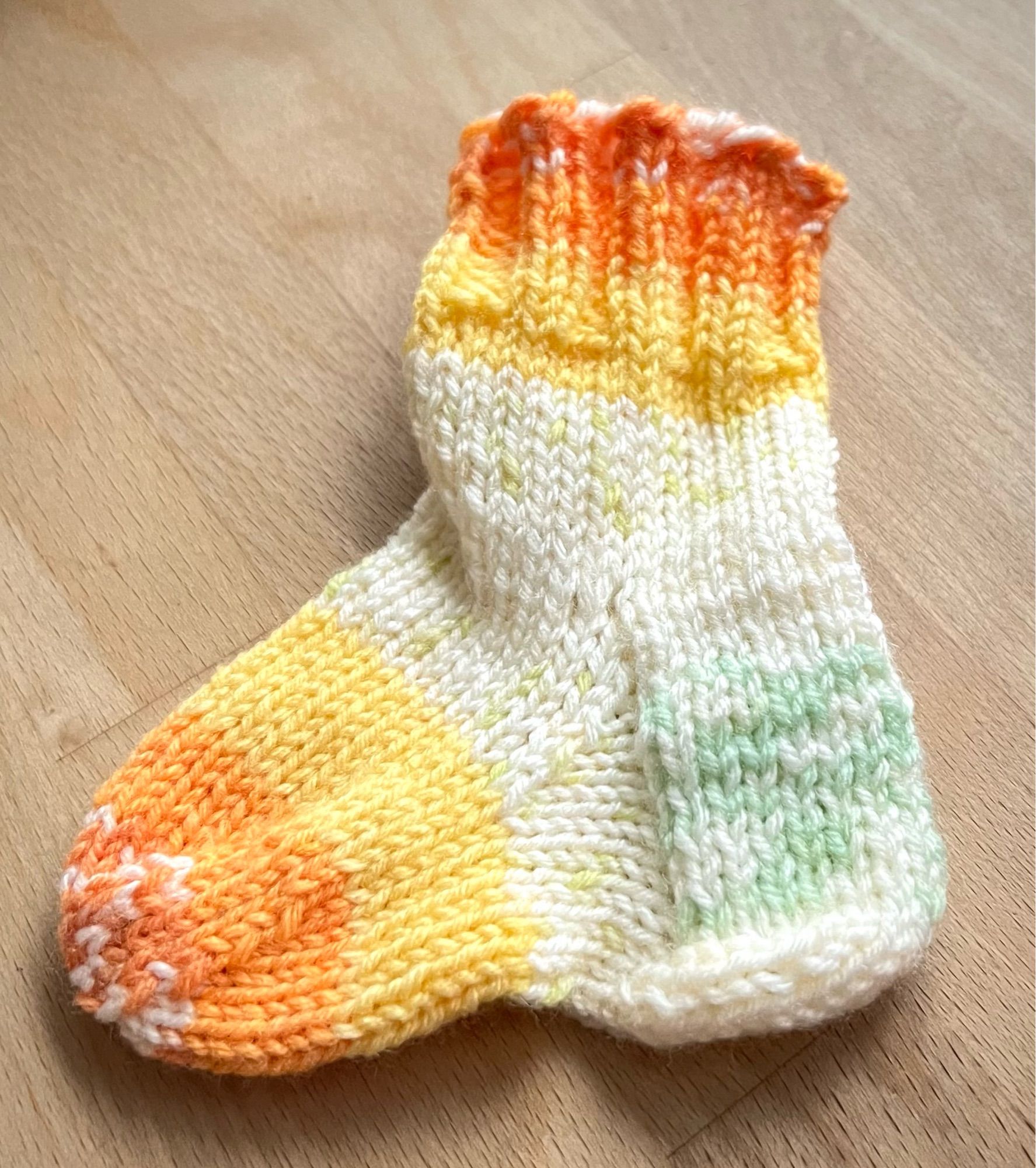 Eine sehr kleine Socke aus Wolle liegt auf einem Holztisch. Verwendete Farben: orange, gelb, weiß sind hellgrün.