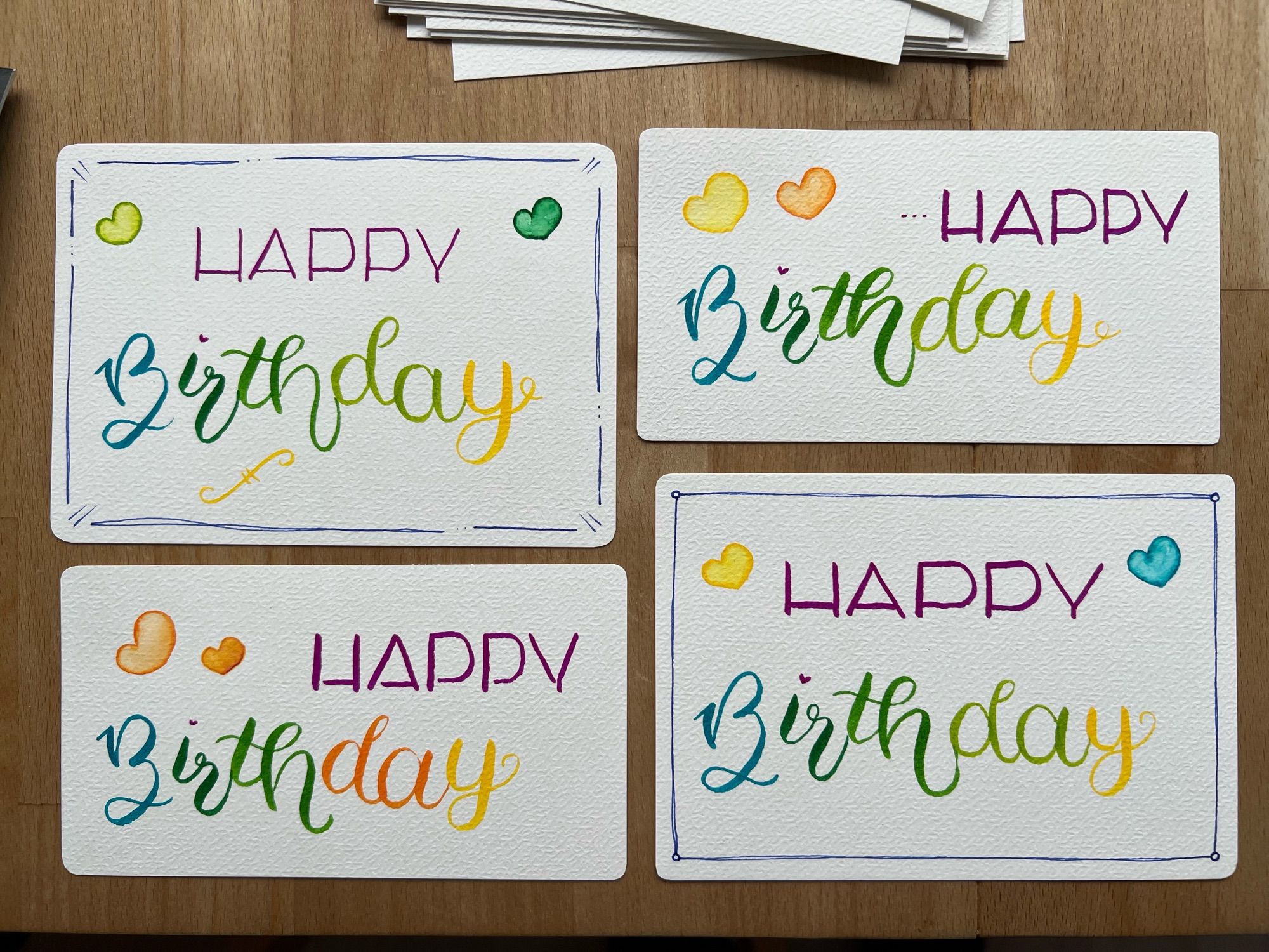 Auf einem Holztisch liegen vier Karten im Querformat. Alle sind weiß und haben abgerundete Ecken. Auf jeder Karte steht „Happy Birthday“. Das Happy ist jeweils in Großbuchstaben und in lila geschrieben. Das Birthday ist jeweils in unterschiedlichen Farben geschrieben. Auf jeder Karte sind zwei bunte Herzen drauf.