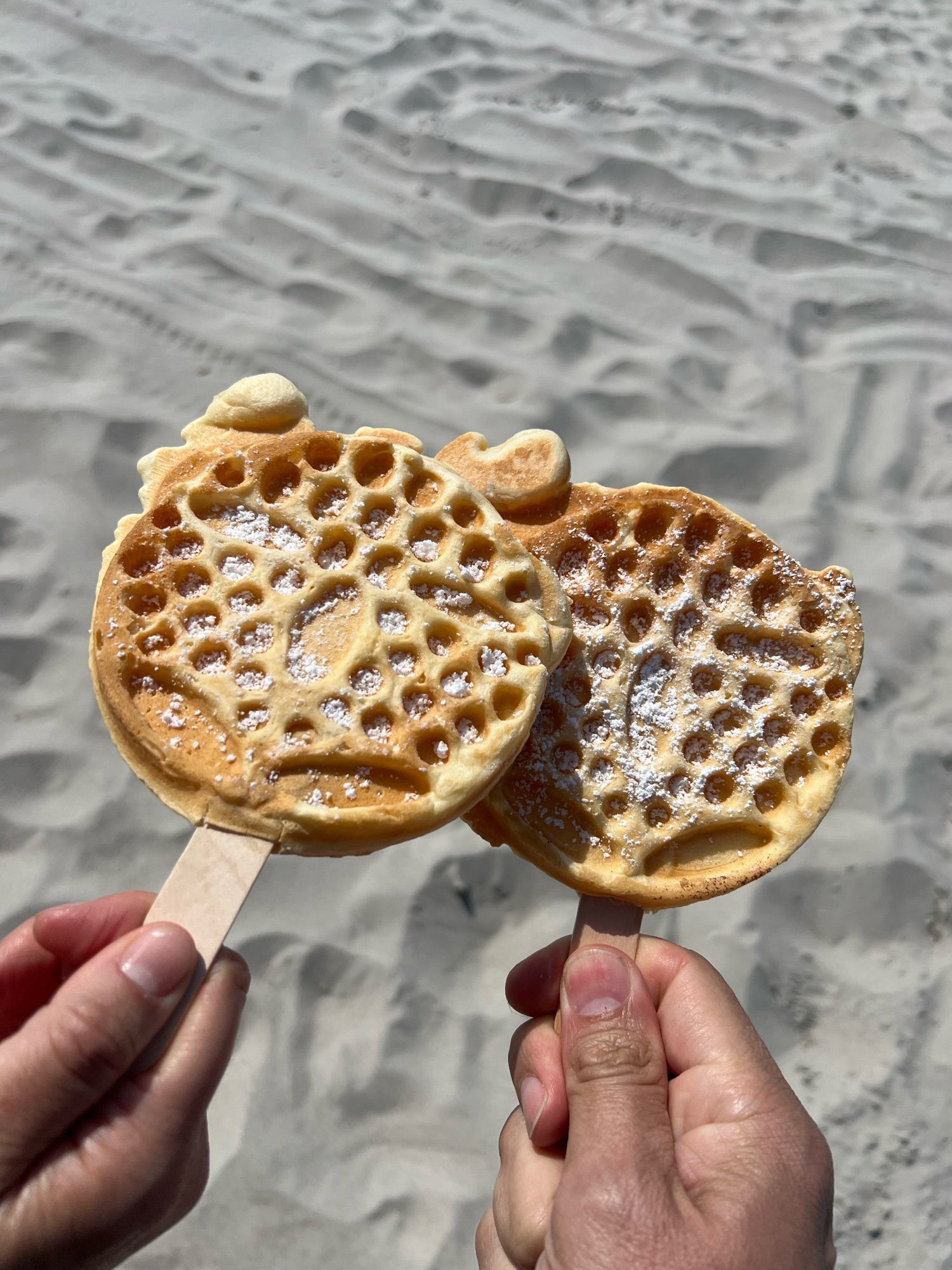 2 runde Waffeln am Stiel, in je einer Hand gehalten. Im Hintergrund der Sandstrand. Es ist sonnig. Auf den Waffeln ist Puderzucker.