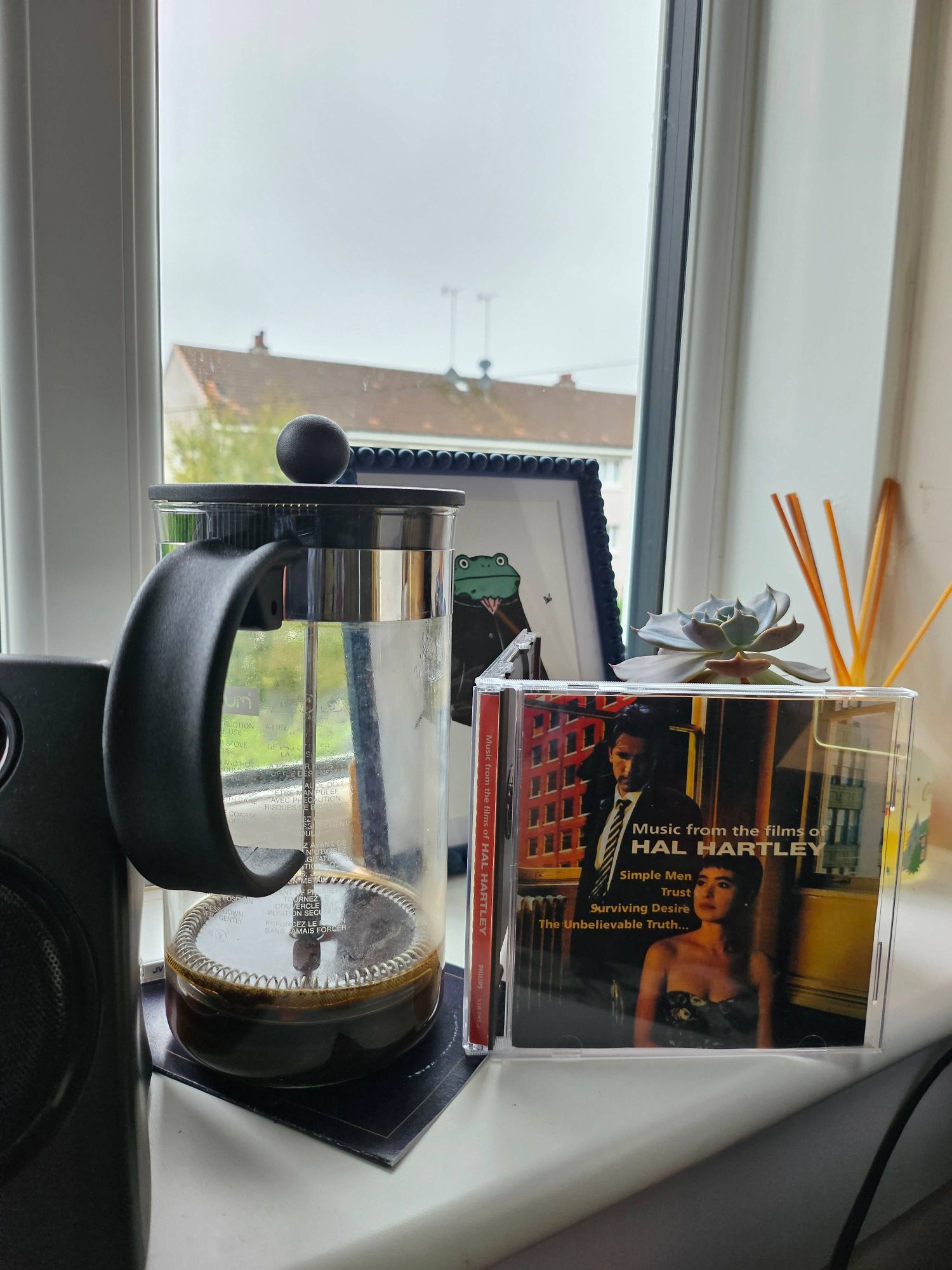 A photo of a crowded windowledge. From left to right we can see: a speaker,  an empty cafetiere, a picture of a frog, a CD (Music from the films of Hal Hartley), a small cactus.