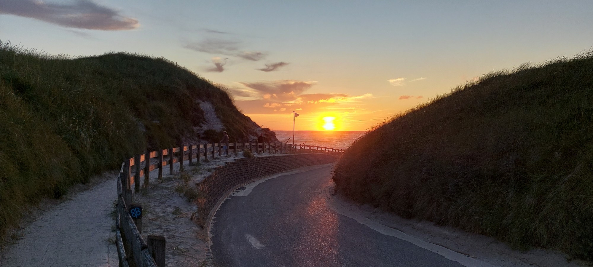 Eine asphaltierte Straße führt zwischen den Dünen durch an den Strand. Die Sonne ist nur noch knapp über dem Meer zu sehen, kurz vor dem Sonnenuntergang.