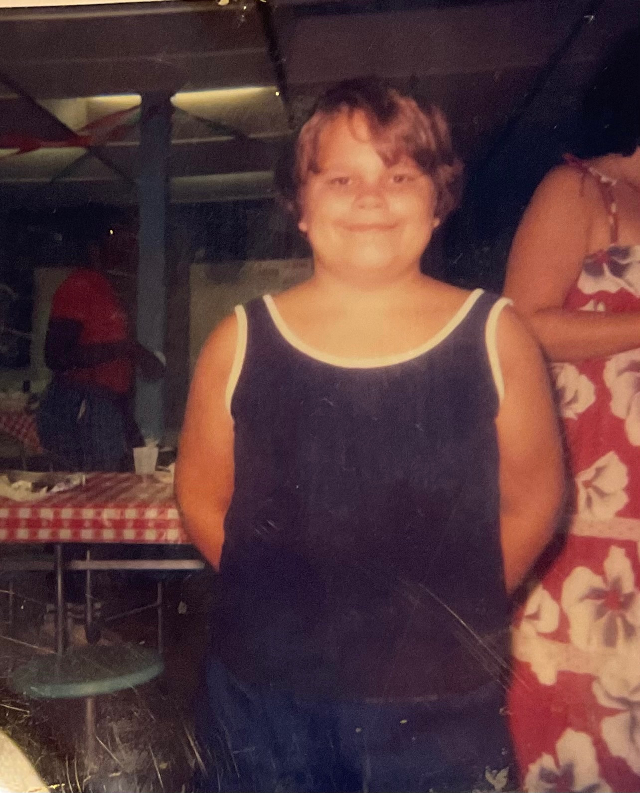 Photograph of a smiling boy (me) wearing a blue muscle shirt and blue shorts with his hands behind his back, standing in an unknown public building’s basement room in front of picnic tables with red and white patterned plastic coverings