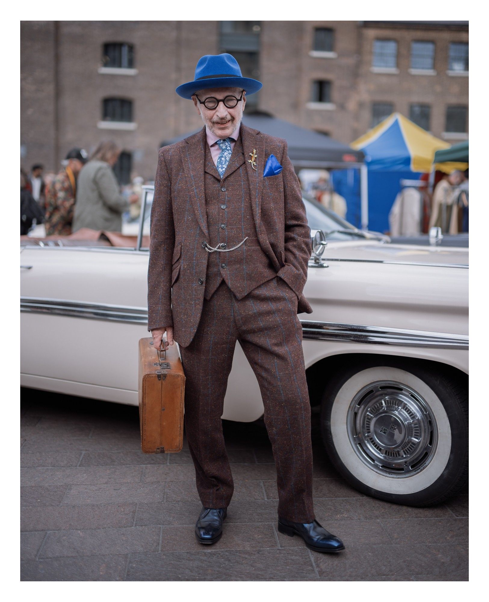 Portrait of a man in vintage clothing carrying a suitcase in front of a classic car.