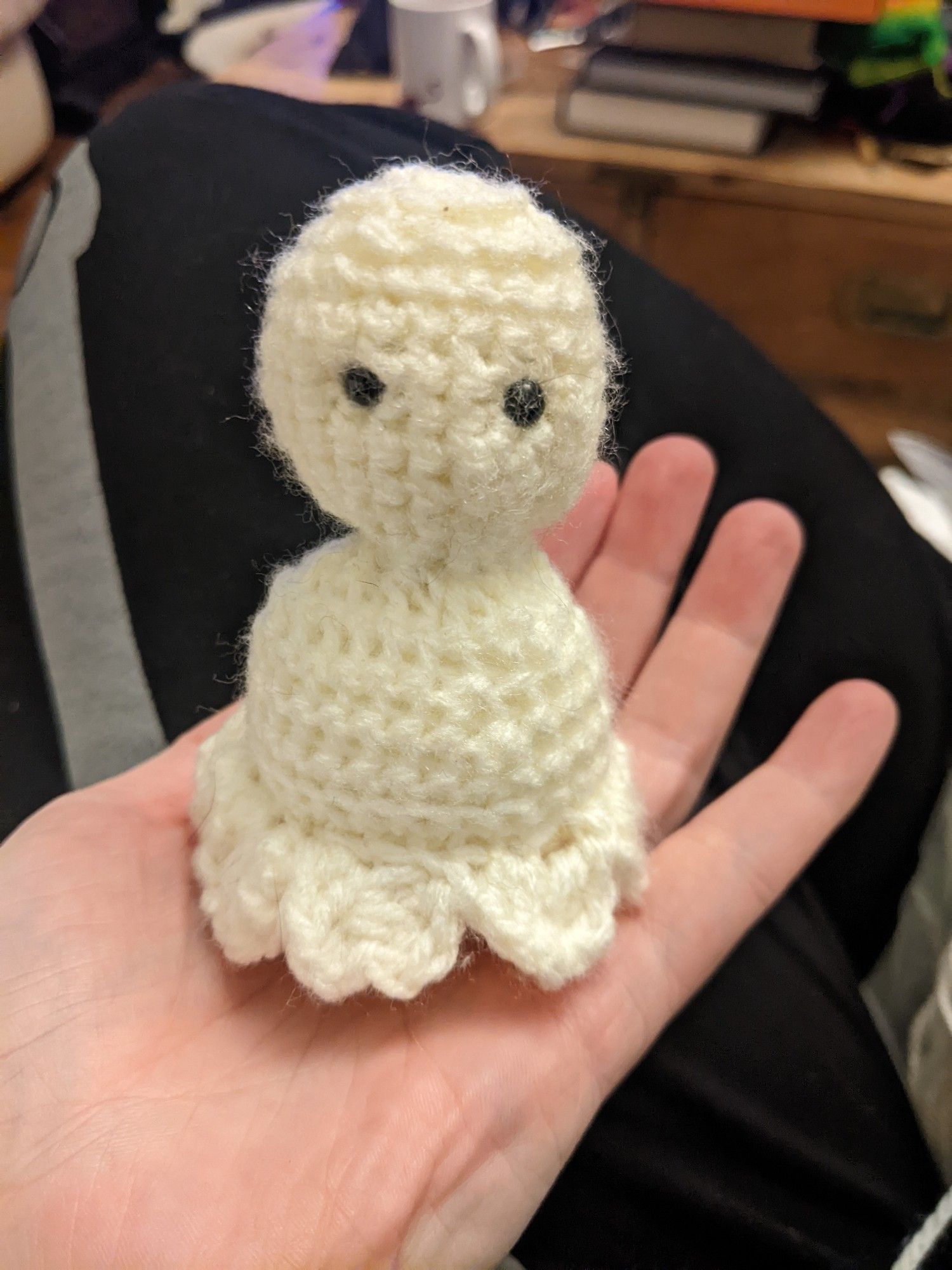 A small crocheted amigurumi ghost made from cream yarn, with small black eyes, sitting in a hand