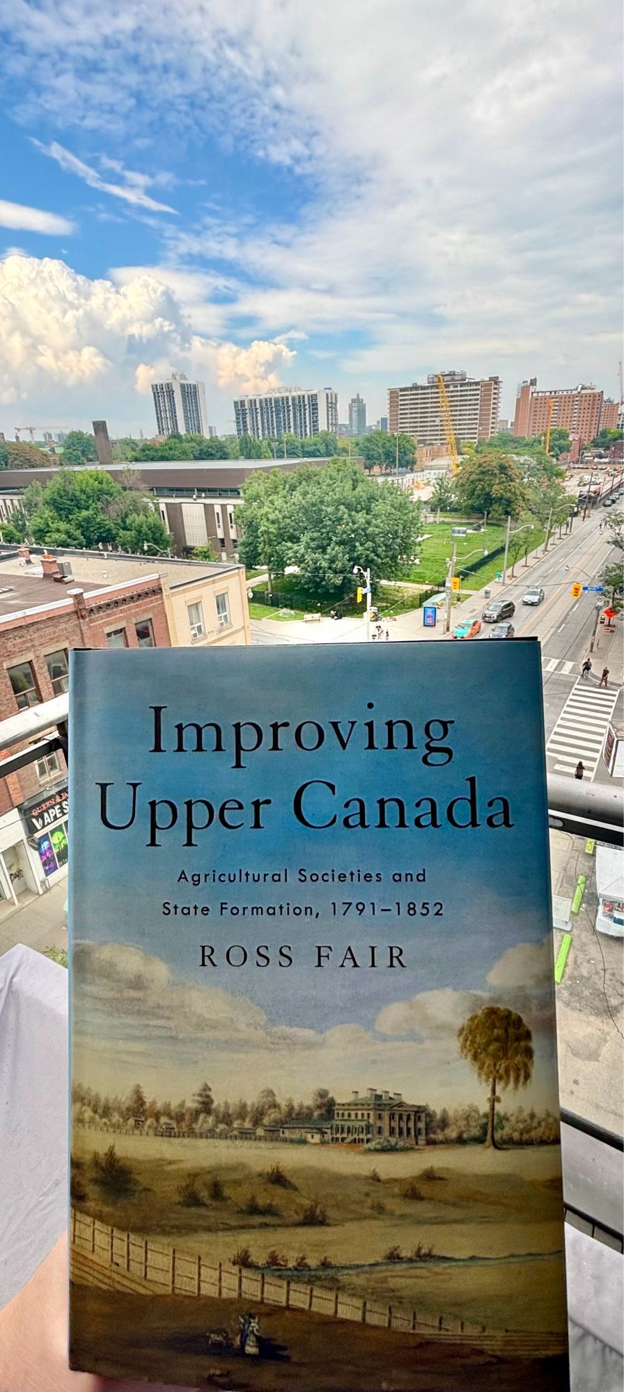 Photo of my book, Improving Upper Canada, that uses John Howard’s 1834 painting of Moss Park estate on its cover. Photo shows book being held today at a similar point of view as painting, with Toronto’s Moss Park neighborhood in background of book 190 years later.