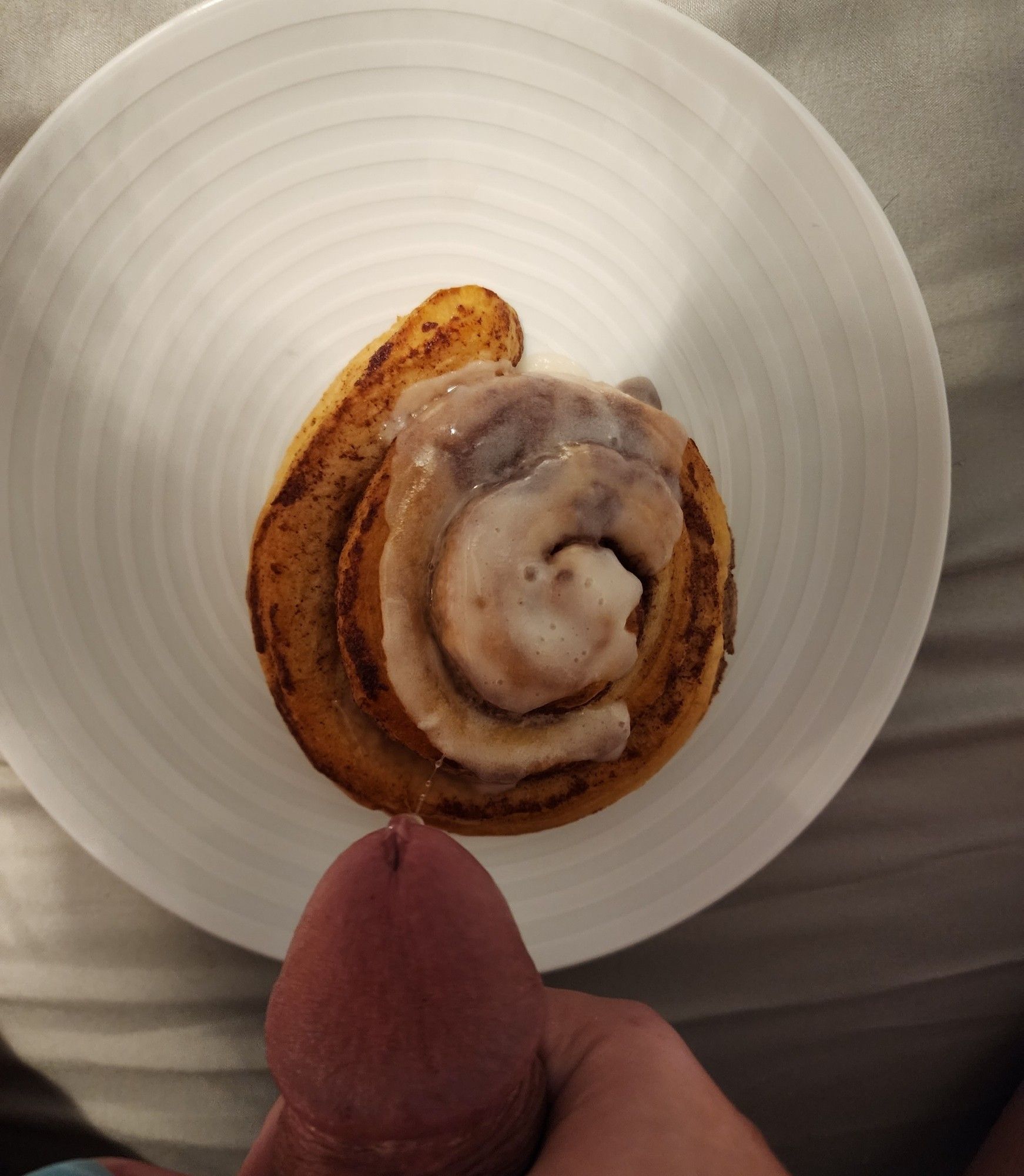 Picture from above. A cinnamon roll on a white place, at the bottom of the picture u can see the tip of junis gock dripping cum onto the cinnamon roll