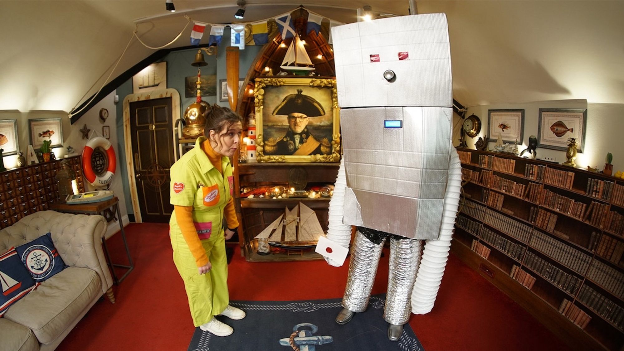 A still from Taskmaster. Rosie Jones in a neon-coloured boiler suit stands next to a shonky-looking, clearly home-made huge cardboard robot, which is really Alex Horne in a cunning disguise.