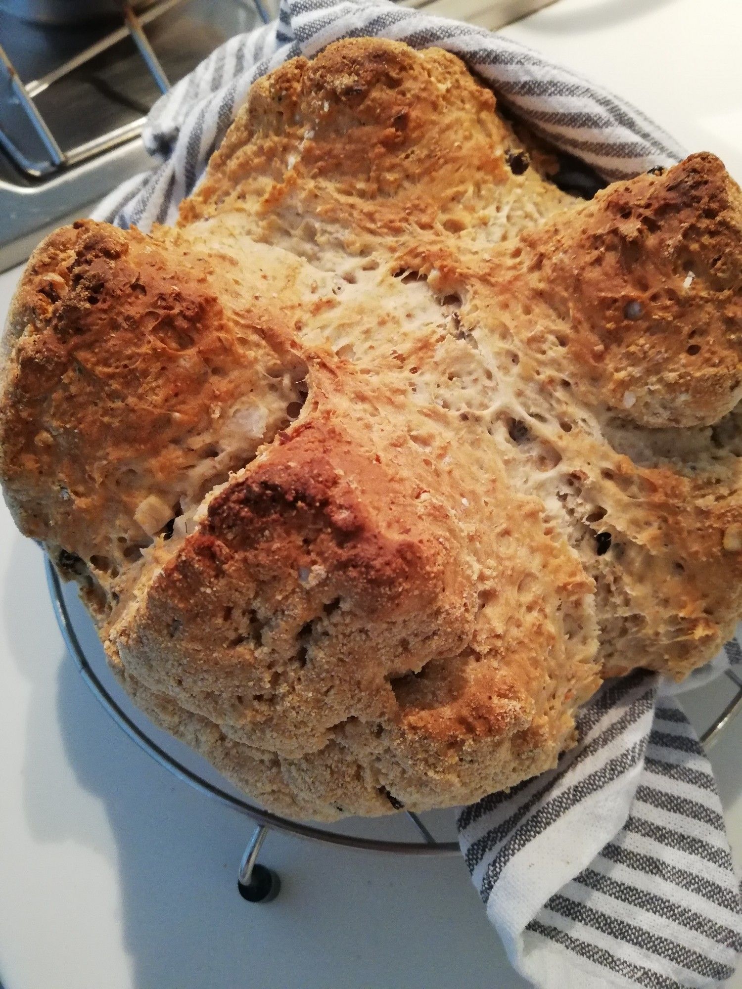 Round cake (loaf) of Irish soda bread partly wrapped in a grey and white tea towel, on a wire rack on a white kitchen surface. Edge of a stainless steel hob visible in top lefthand corner