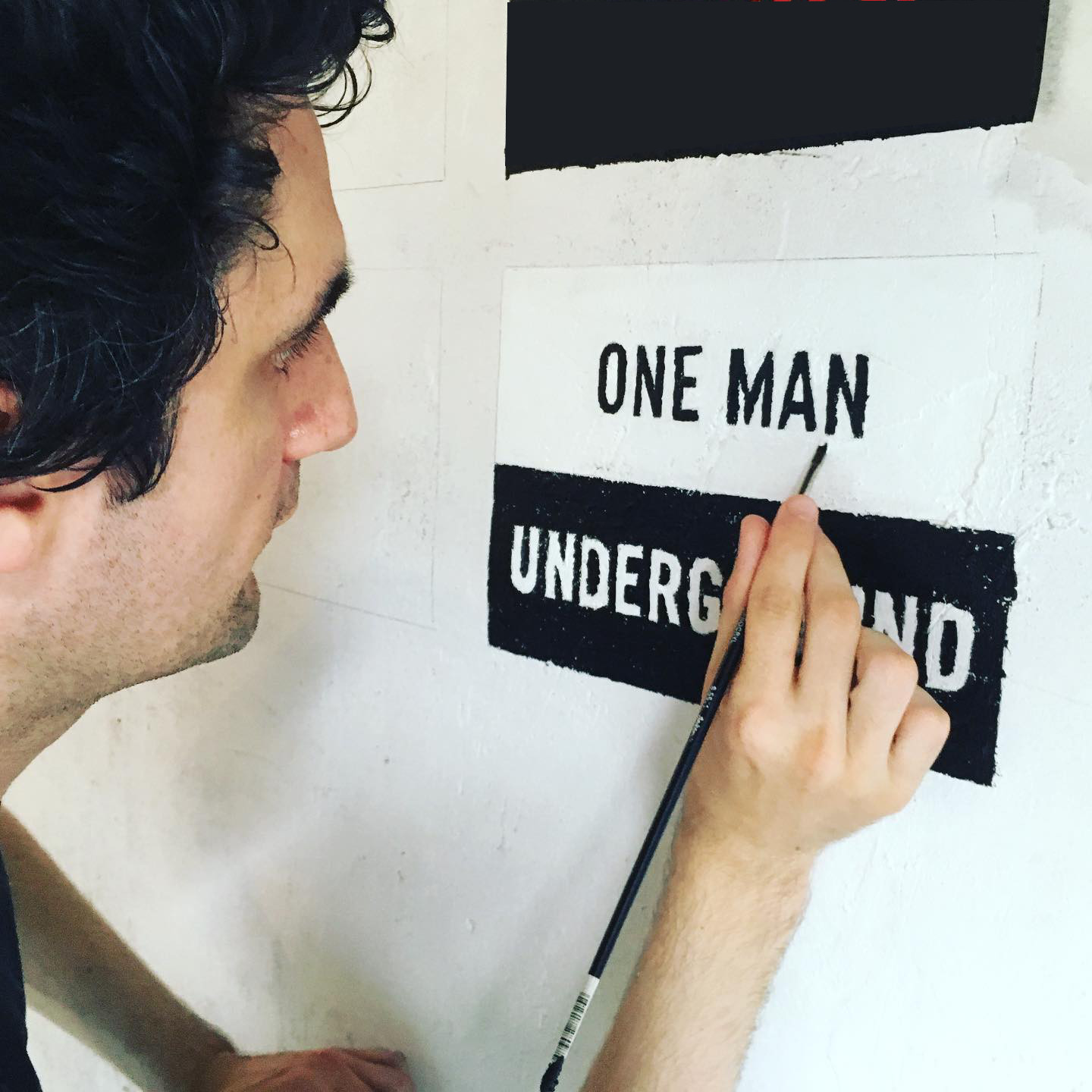 A close up of a white man with big, curly black hair and stubble painting the words, "ONE MAN UNDERGROUND," onto a white wall.