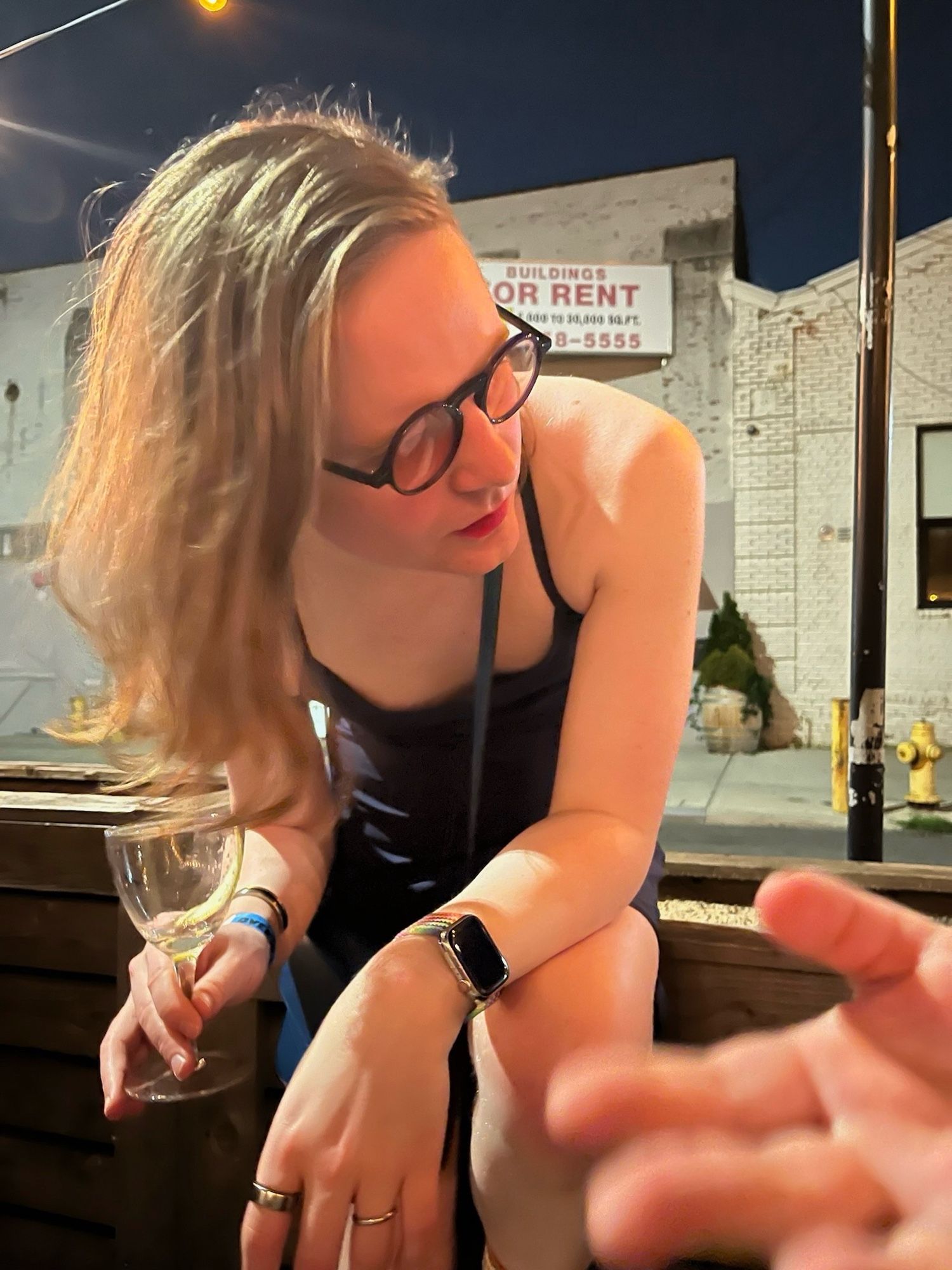 mb, a white nonbinary transfem, in a blue dress, red lip, and blue glasses, sitting on an outdoor dining structure, with a nearly empty martini in hand outside the Gender Reveal lives how afterparty in Gowanus.