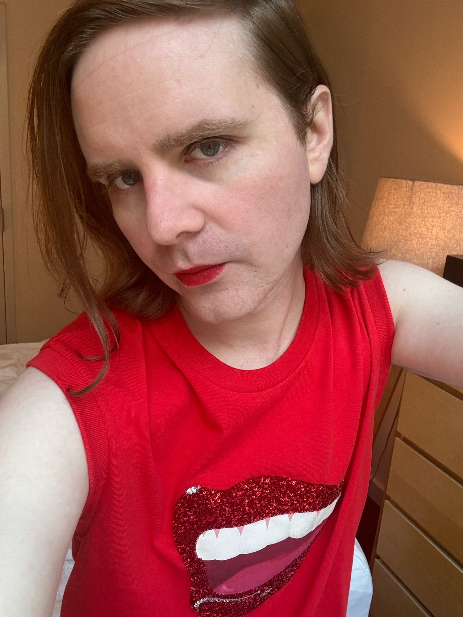 mb, a white nonbinary transfem, in her room wearing a loud red shirt with a sparkly mouth and red lipstick.