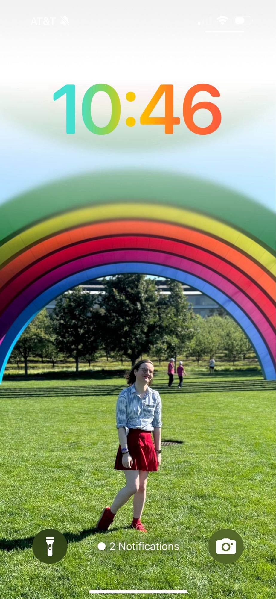 mb, a white nonbinary transfem under the rainbow stage on the grass at Apple Park in a red skirt and blue striped shirt.