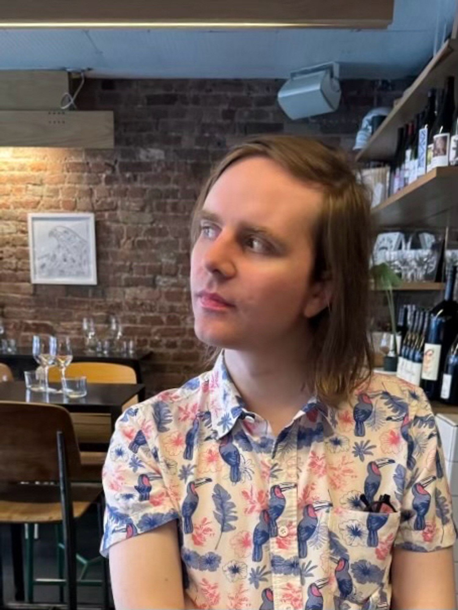 mb, a white nonbinary transfem, looking pensive at a downtown wine bar in a pink and blue toucan-print shirt.