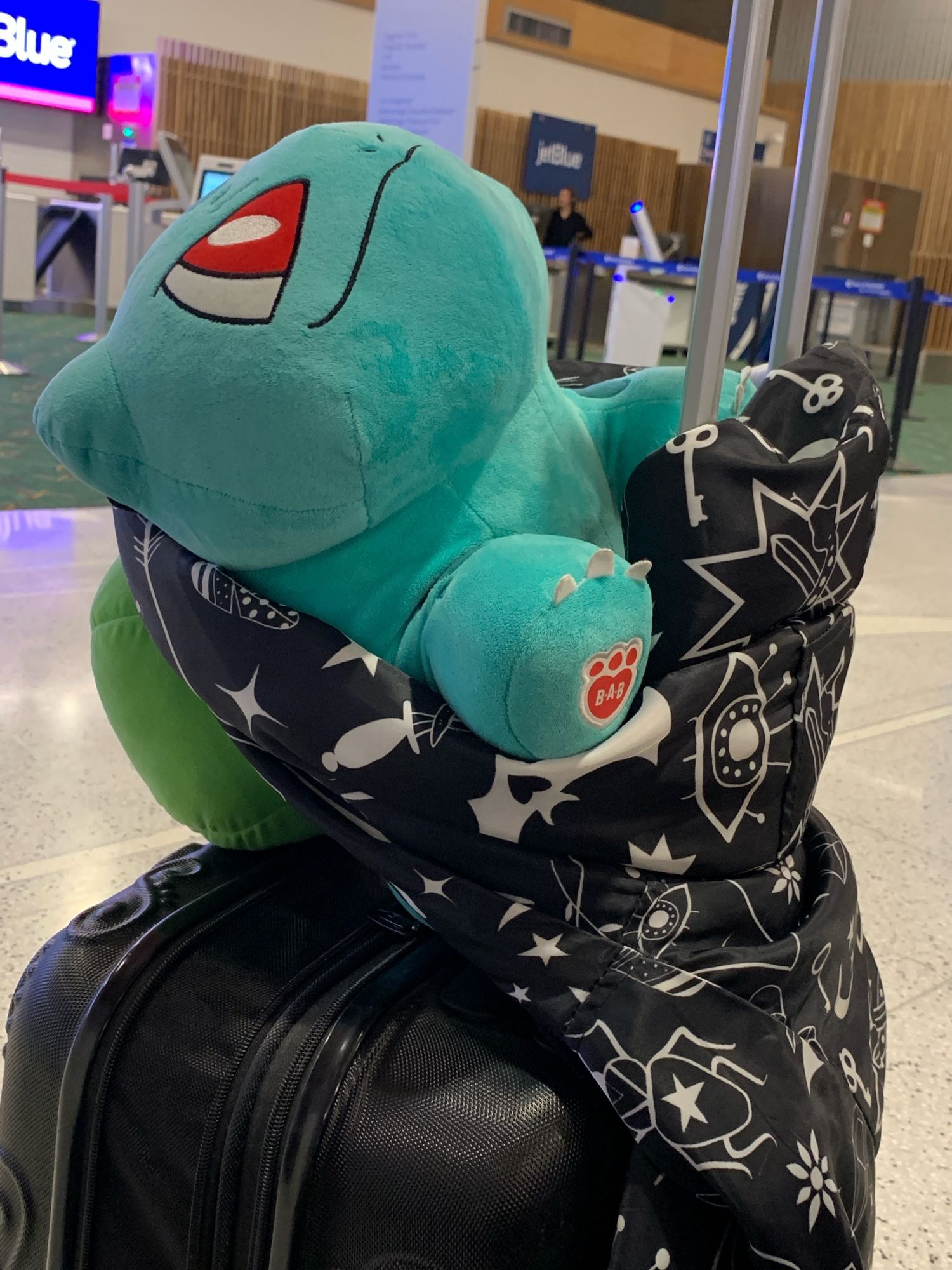 Image of a plush bulbasaur attached to a suitcase handle by a tied jacket.