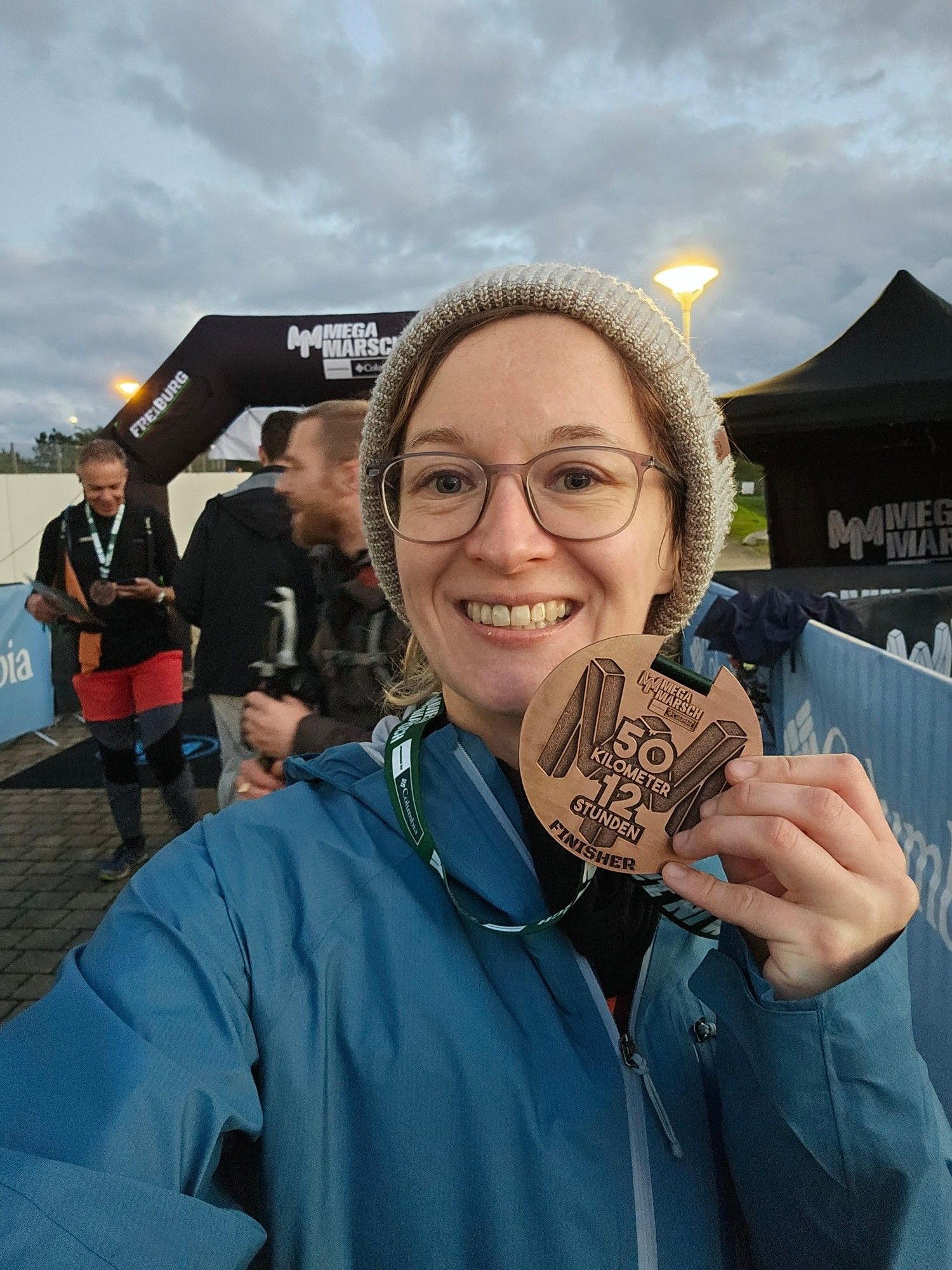 Ich, mit Regenjacke und Wollmütze, halte meine Finishermedaille in die Kamera.