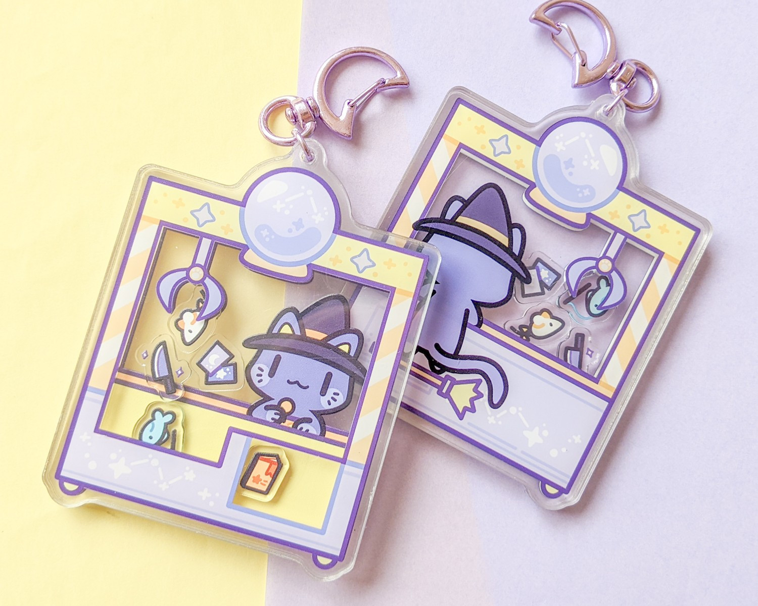 Acrylic shaker charm of a witchy cat at a UFO catcher machine.  The shakable pieces inside the machine are of a toy mouse, tarot cards, toy fish on a string, book, and knife.  The charm is on a purple and yellow background.