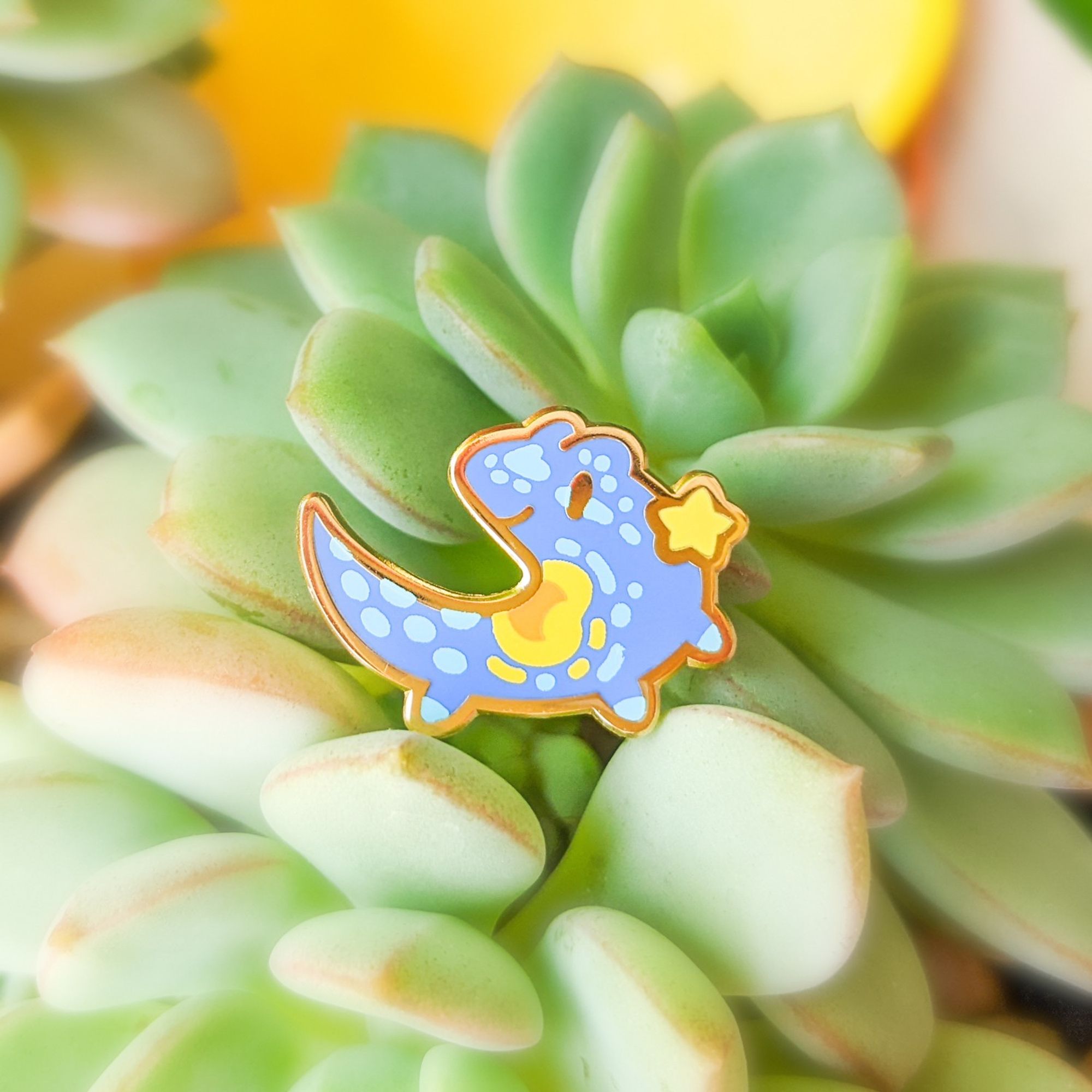 Photo of an enamel pin of a blue gecko with a little star.  The background is a succulent.  The gecko is the Van Gogh Starry Gecko.