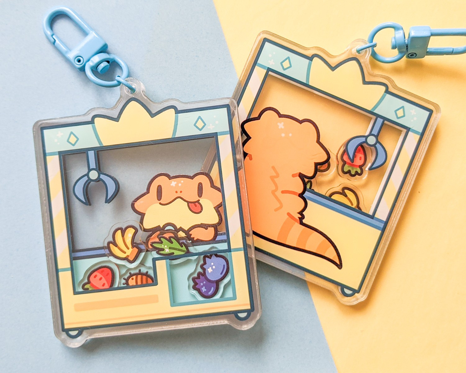 Acrylic shaker charm of a bearded dragon at a UFO catcher machine.  The shakable pieces inside the machine are of a strawberry, banana, blueberry, arugula, and roach.  The charm is on a blue and yellow background.