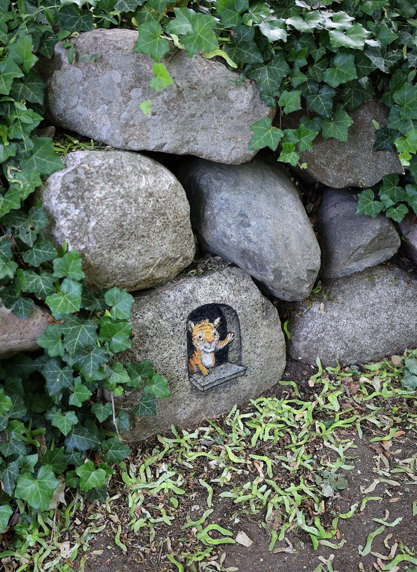 Zeichnung auf einem Stein am Wegrand. Mehrere grosse Steine sind daneben und darüber. Sie sind von Efeu überwachsen.
Die Zeichnung simuliert ein Fenster im Stein, mit Fenstersims. Ein kleiner Tiger schaut heraus.