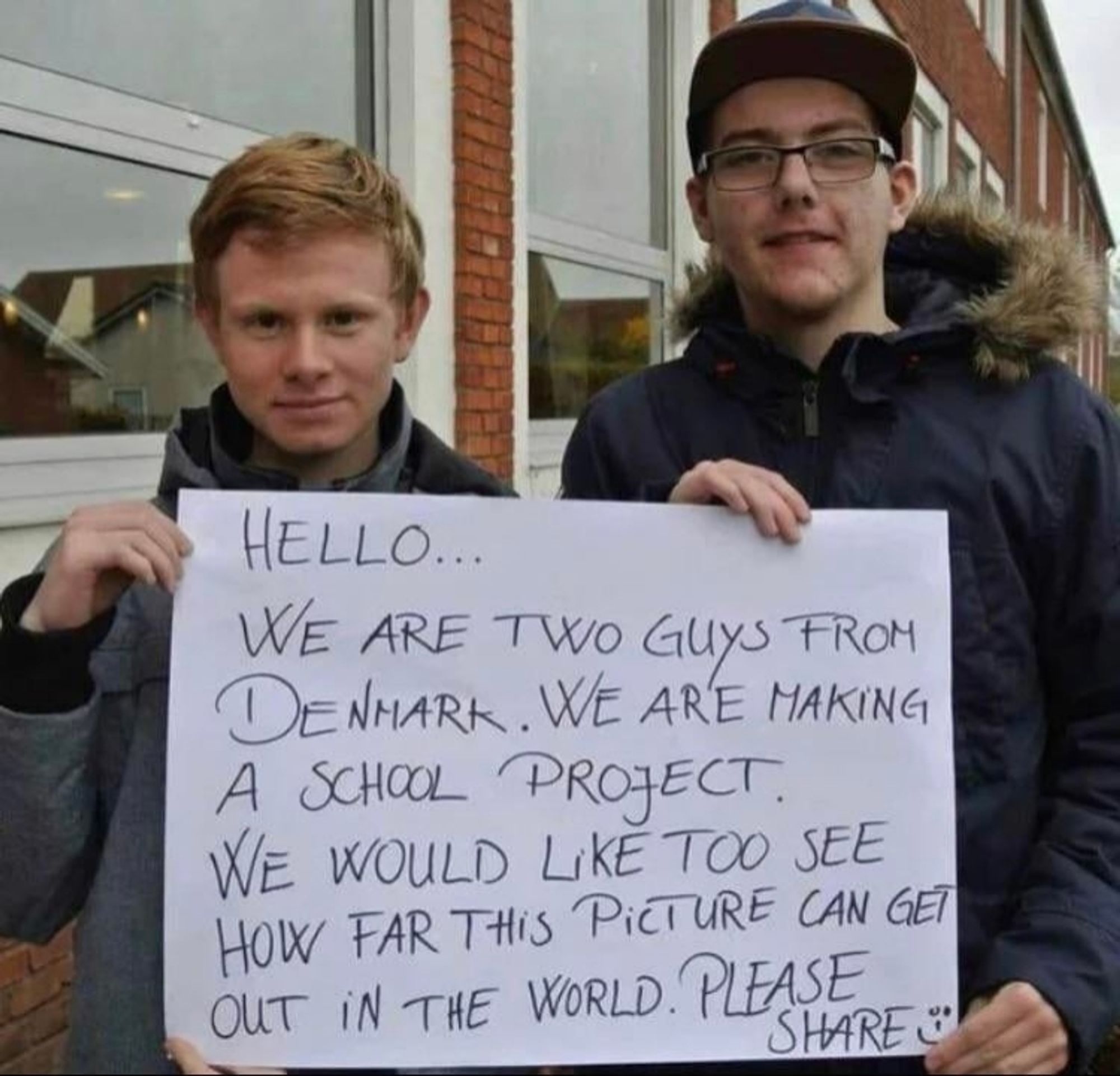 Zwei junge Menschen halten ein Plakat mit der Aufschrift " Hello, we are two guys from Denmark. We are making a school project.we would like to see how far this picture can get out in the world. Please share."