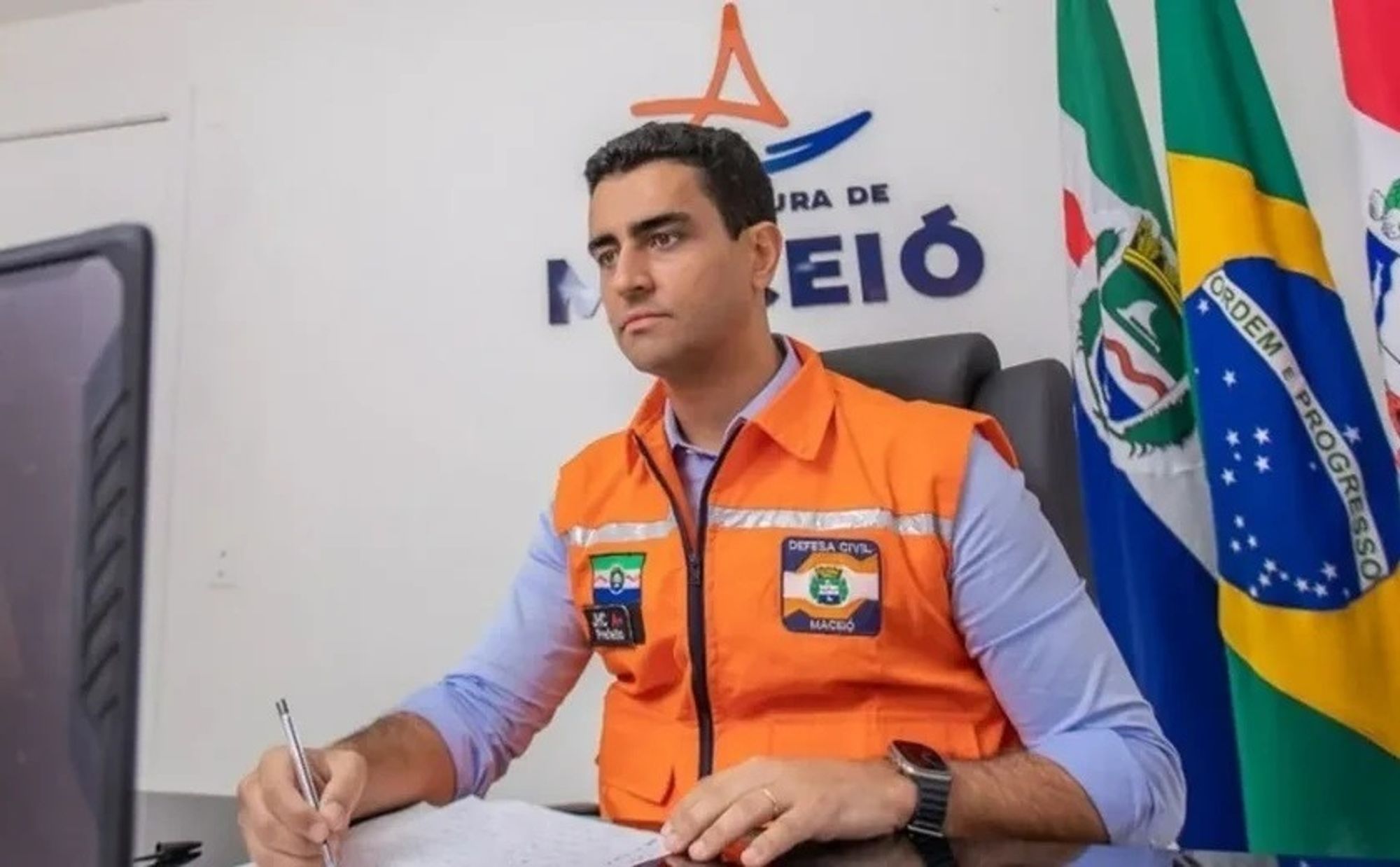 Prefeito de Maceió, JHC, sentado na frente de um computador usando camisa social azul e um colete laranja da defesa civil. Ele segura uma caneta e parece escrever algo num papel. Ao fundo, um desenho de uma jangada e "prefeitura de Maceió" parcialmente cobertos pela cabeça dele. Ao lado, bandeiras de Maceió, do Brasil, e de Alagoas.