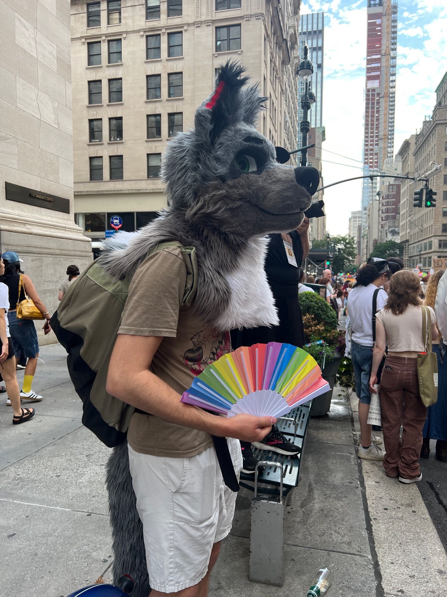 A gray wolf partial fursuiter fans a rainbow fan in NYC