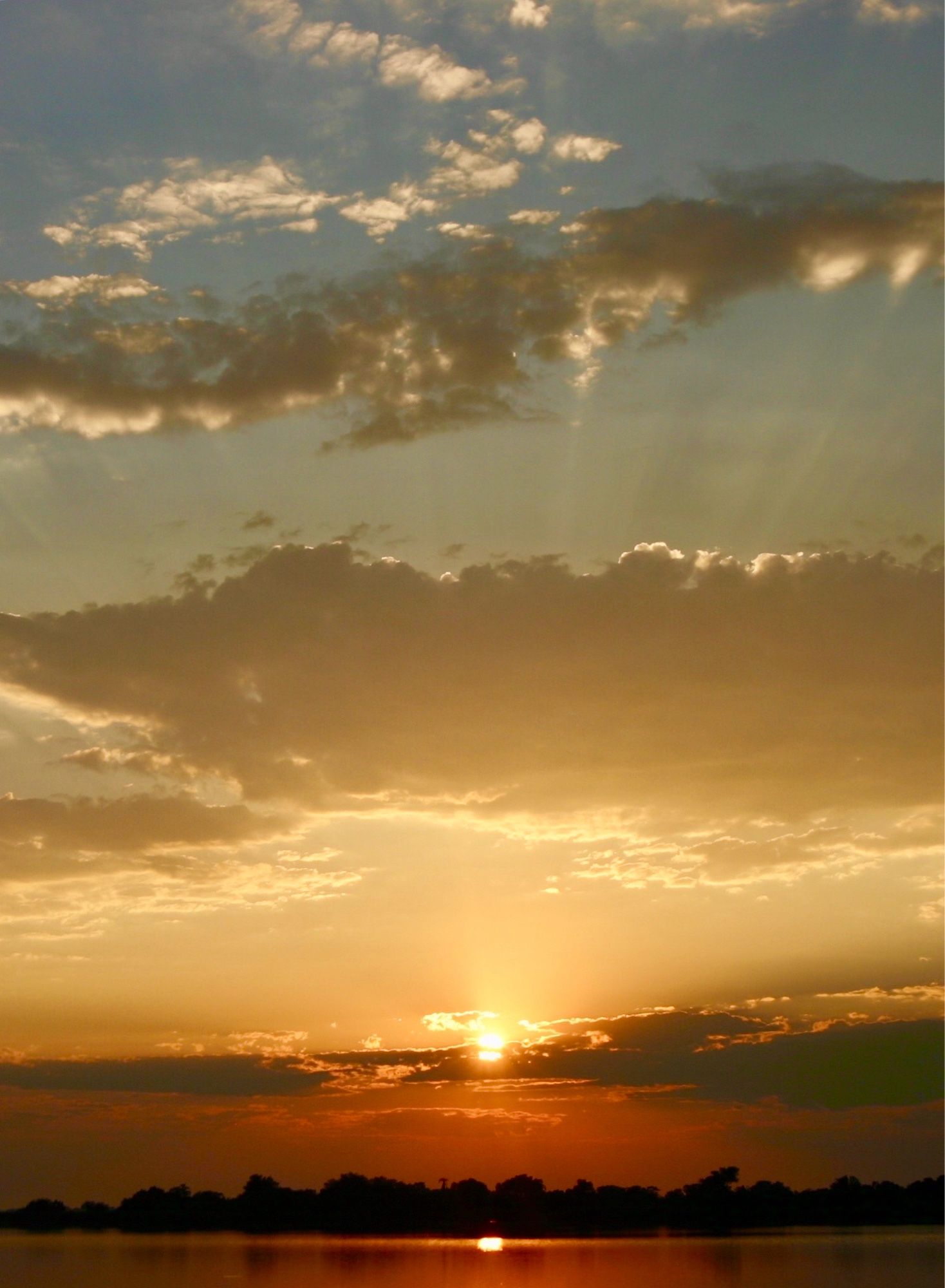 Sun has risen for a new day on the Okavango Delta.

A lake, on the far shoreline bounded by silhouettes of tree, reflects the early morning sun red whilst the sun itself is above some low lying clouds on the horizon.  The sky above shows some of the sunlight as rays appearing from a higher cloud

Okavango Delta, Botswana