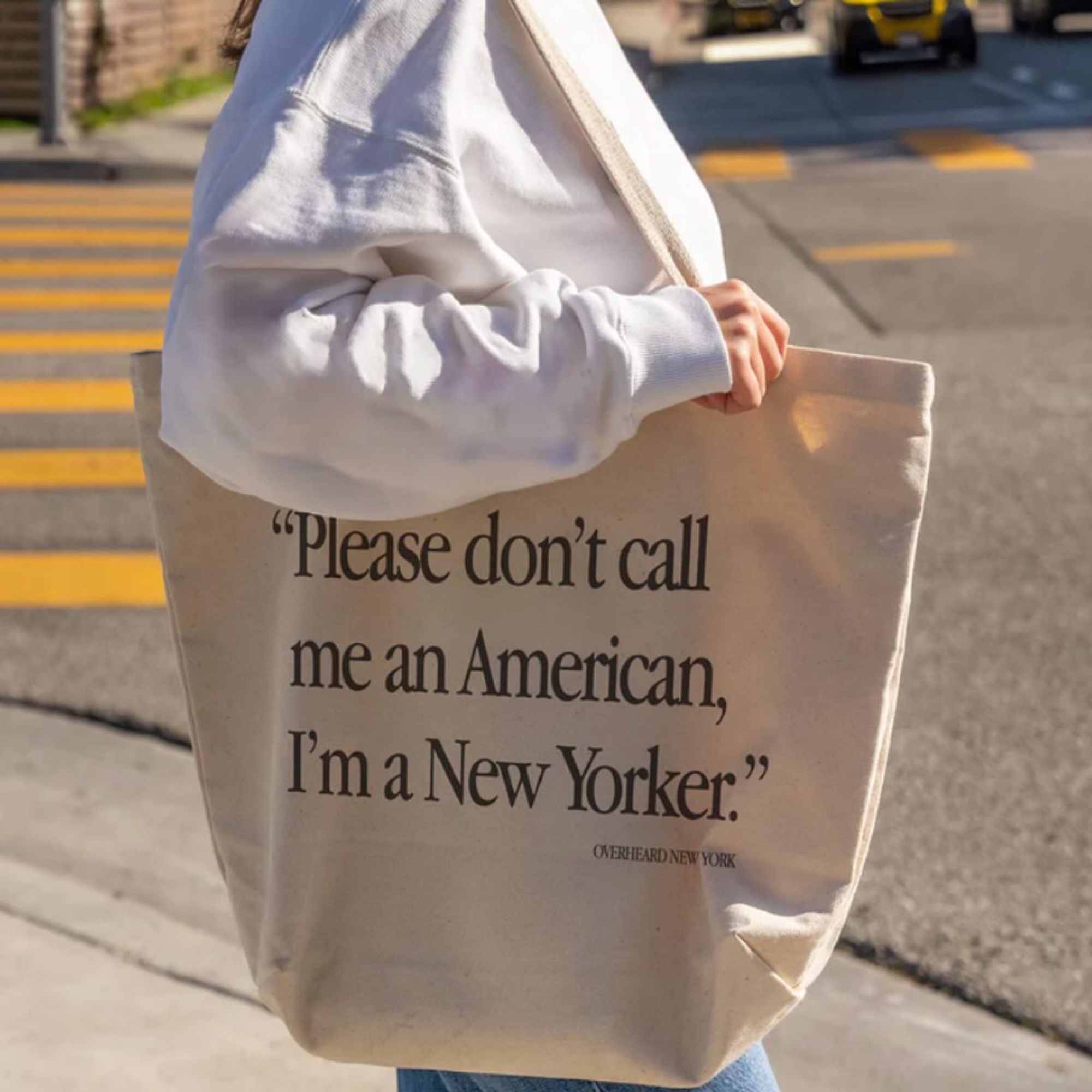 a woman standing on an NYC street with a tote bag with the quote "please don't call me an american, i'm a new yorker"