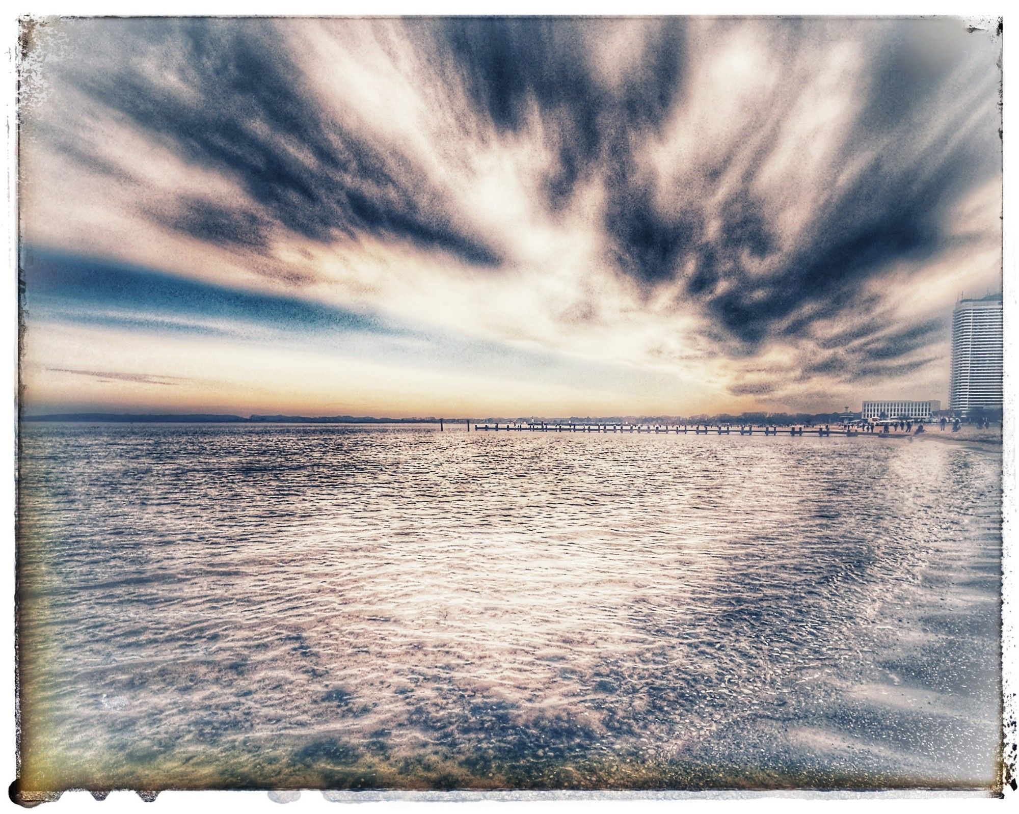 Am rechten Bildrand steht das Maritim an der Promenade in Travemünde, dahinter geht die Sonne unter und verleiht dem mittigen Horizont über der Ostsee einen orangenen Streifen und den Wolken am Himmel etwas apricot.