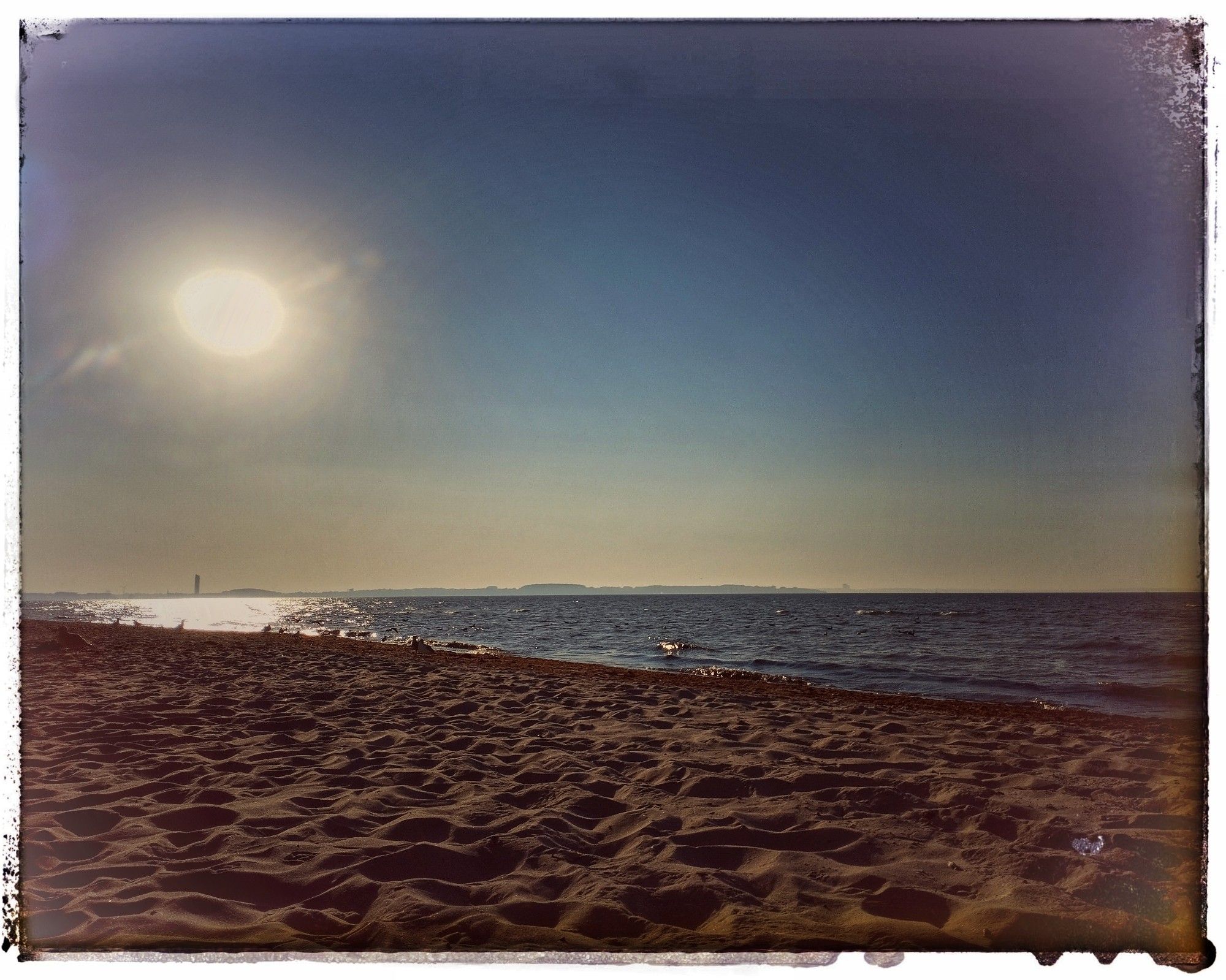 OstseeStrand im herbstlichen Sonnendunst. Ein Streifen Meer unter blauem Himmel, der Horizont milchig gelb.