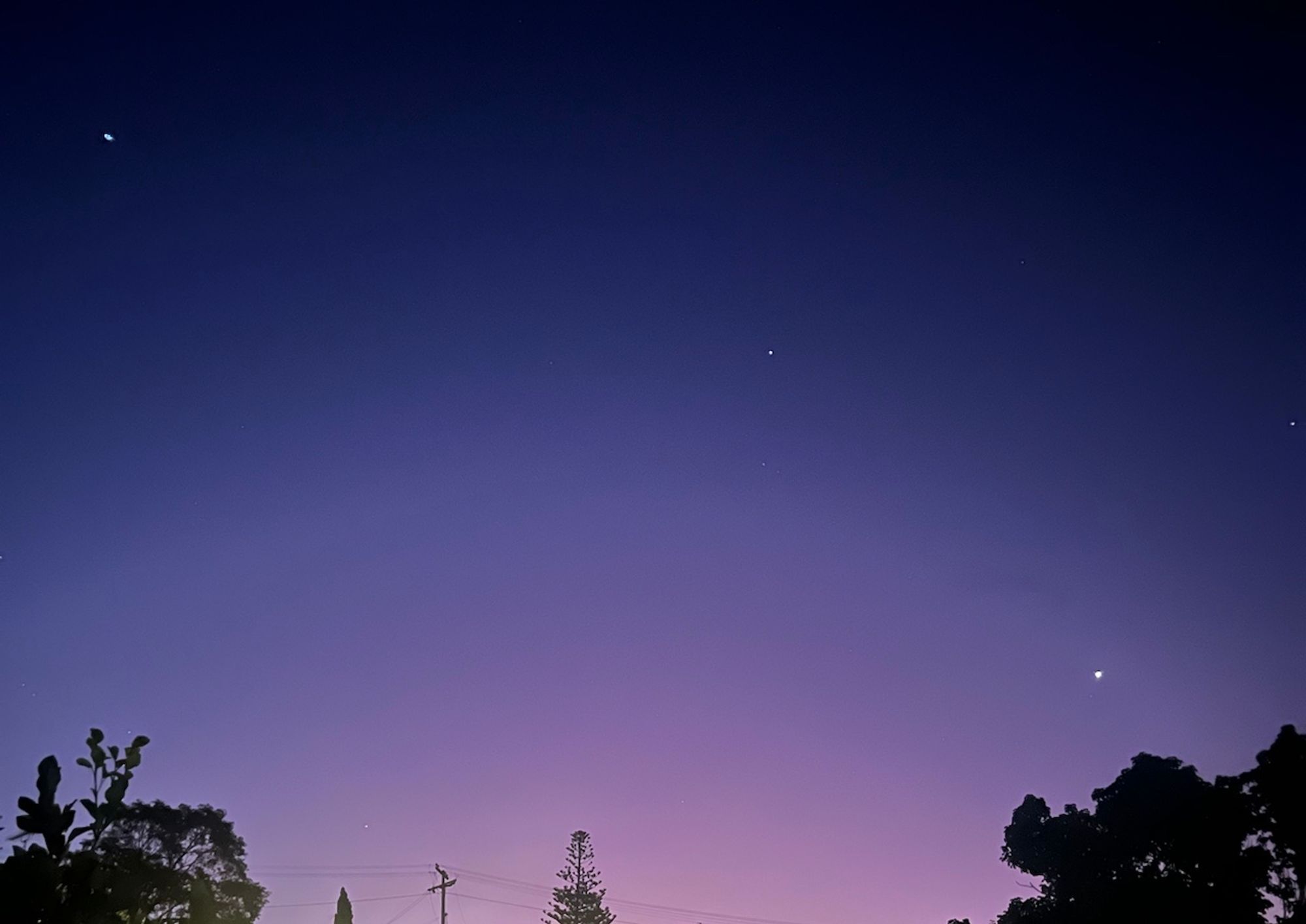 A lovely blue gradient sky, at night, with stars. There are trees in the bottom left and right of the image. The light from my neighbors clashes with the blue gradient to make an even more powerful image.