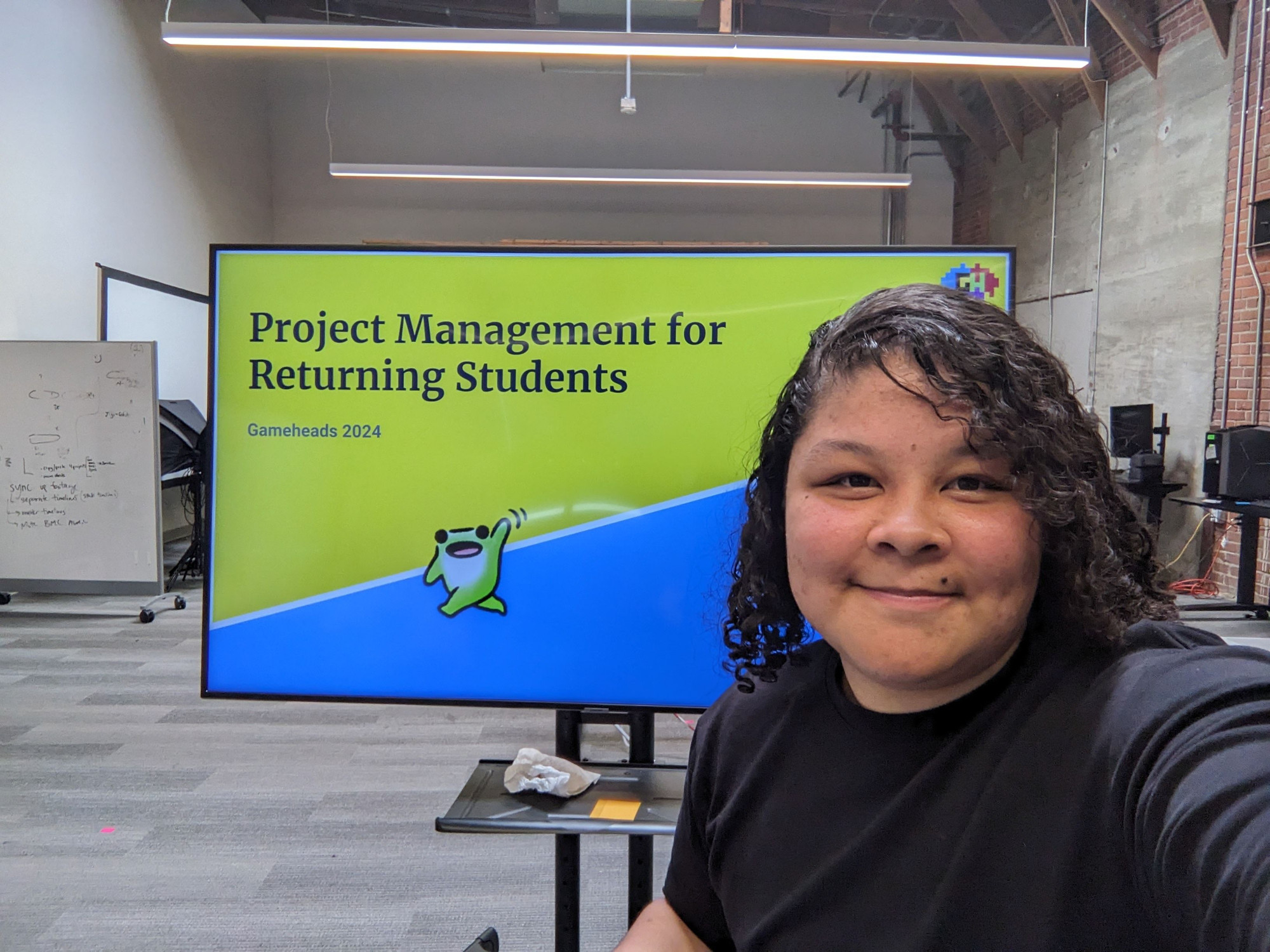 Yellow and blue slide deck on a large TV with the title "Project Management for Returning Students" for the summer 2024 Gameheads cohort. NyameDev holding the camera for a selfie before the talk!