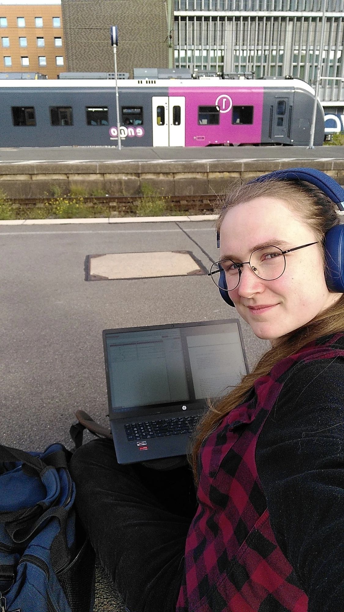 Me (very long hair, black and red clothing) sitting on the ground at a train station, two bags next to me and a laptop on my legs. Two windows with text are open on the screen. Tracks and a train in the background. It's sunny and I'm wearing headphones.