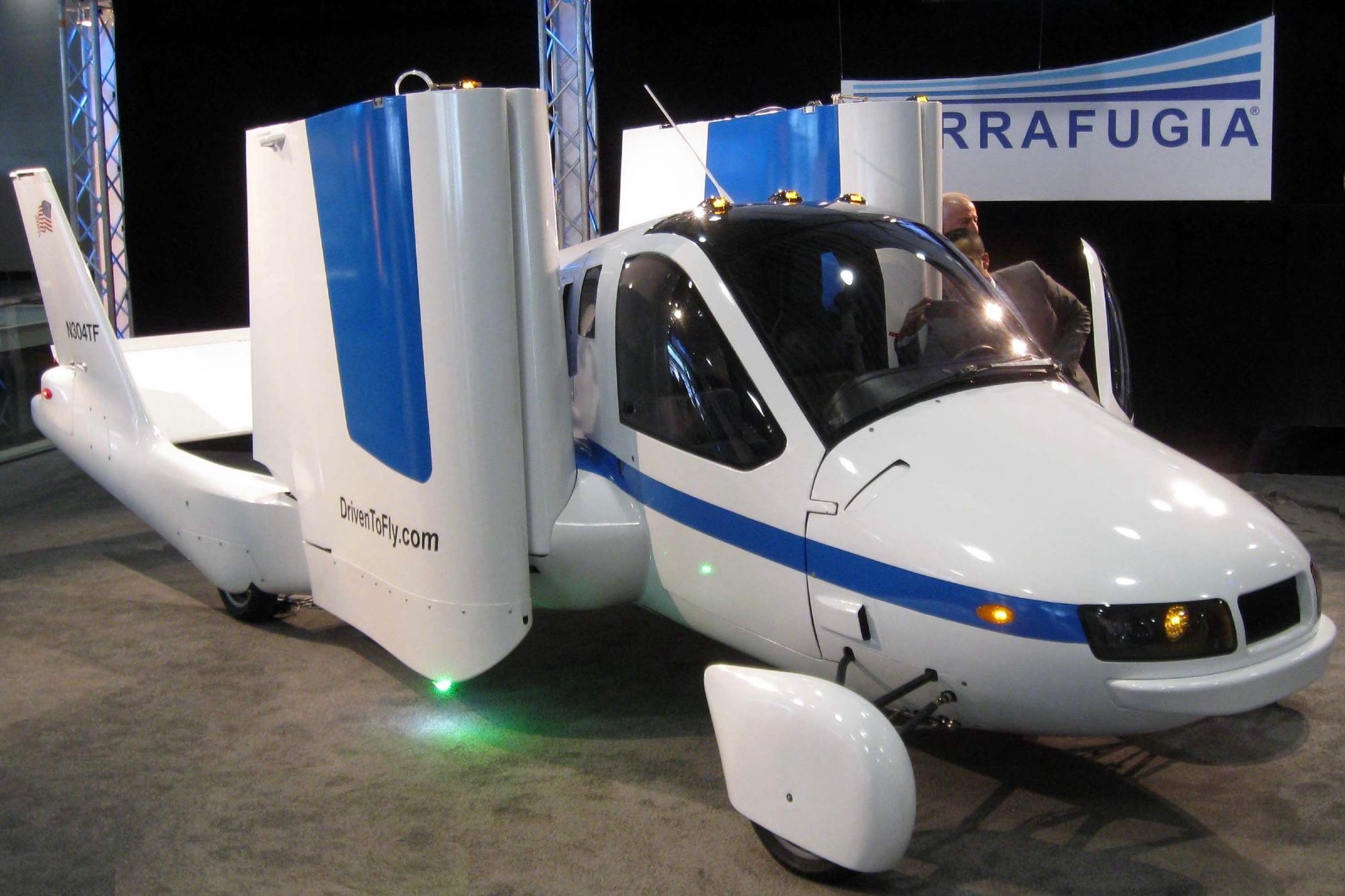 A picture of the flying car which is the subject of this post. It looks like a fairly conventional vehicle, mostly white with blue accent stripes, with wings folded alongside the main body. Vertical and horizontal stabilisers are also visible. The website « DriveToFly.com » is painted on the right, folded wing. This photo was taken at the 2012 New York International Auto Show. (📸 M. Warren via Wikimedia under CC BY-SA 3.0)