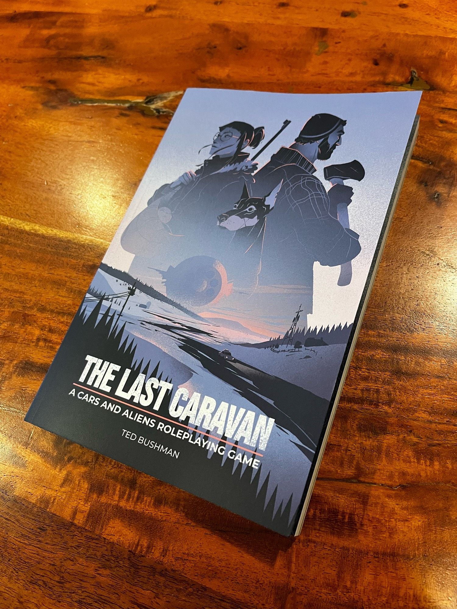 A test print of The Last Caravan by Ted Bushman resting on a wooden table. The cover depicts a rural highway blanketed with snow, a lone car driving toward us--away from an ominous alien spaceship on the horizon. Three travelers stand side by side in the sky: a woman with a baby and a sniper rifle, a man with a woodcutting axe, and a resolute dobermann pinscher.

The Last Caravan: A cars and aliens roleplaying game