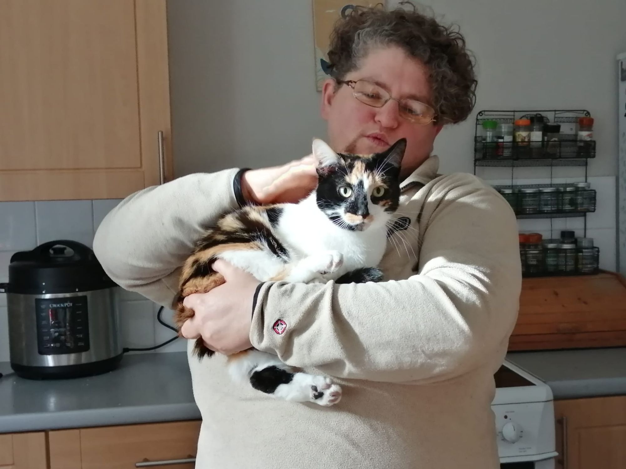 A white perusing with short curly hair holds a tortoiseshell and white (calico) cat  who is looking at the camera.  There are kitchen objects behind