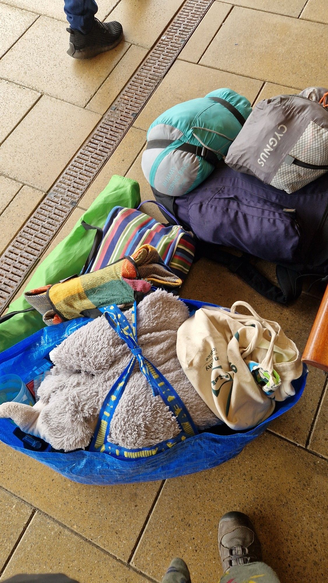 A heap of luggage on a train platform.  There is a rucksack with attached tent,  sleeping bag and stripy bag with wellies; A green camping chair; and IKEA blue bag with attached blanket and an elephant in the top.