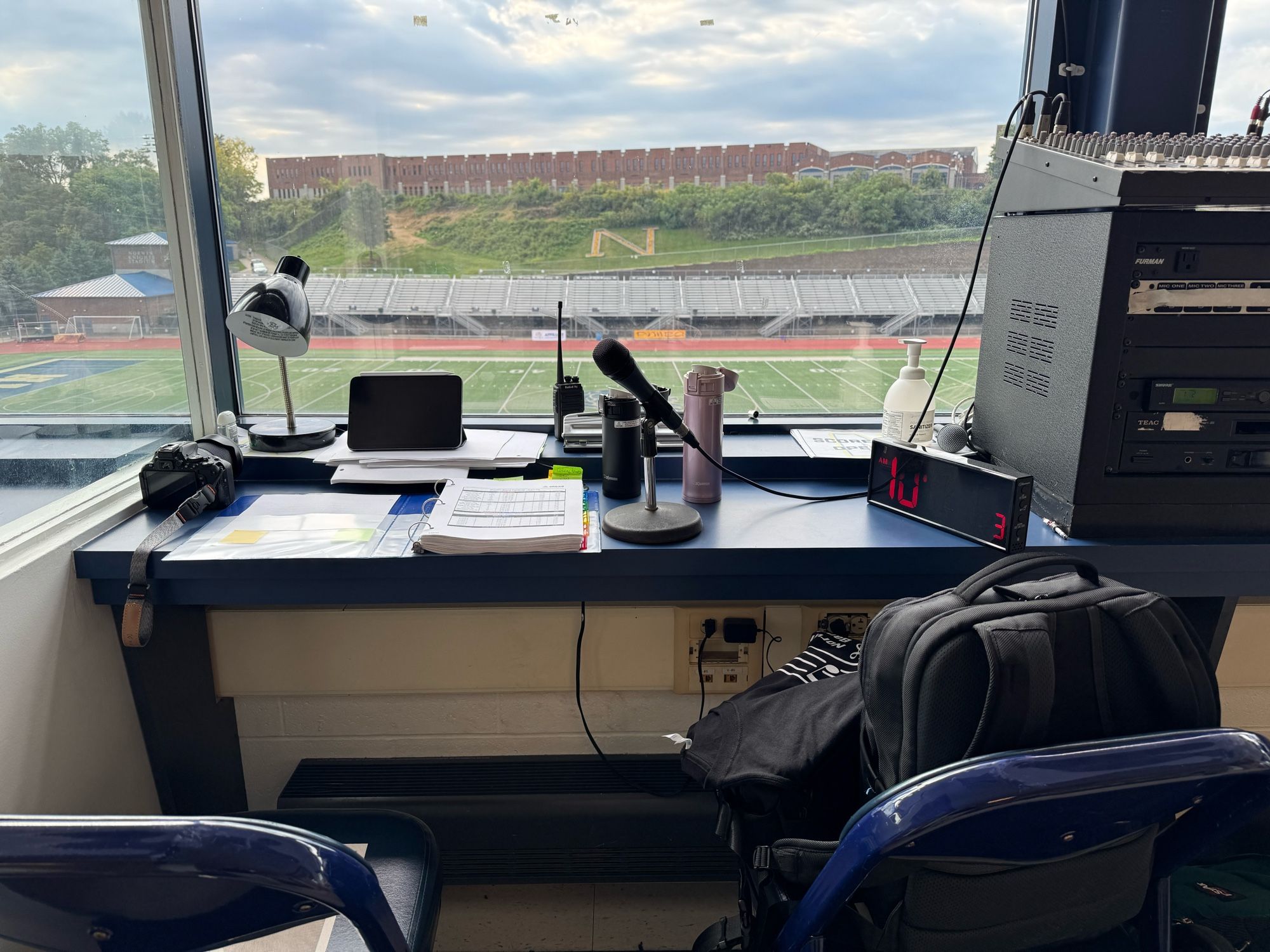 A view from a press box showing a desk with a lamp, microphone, camera, and a digital clock. In the background, a sports field is visible with bleachers and a hill. There is also a backpack on a chair.