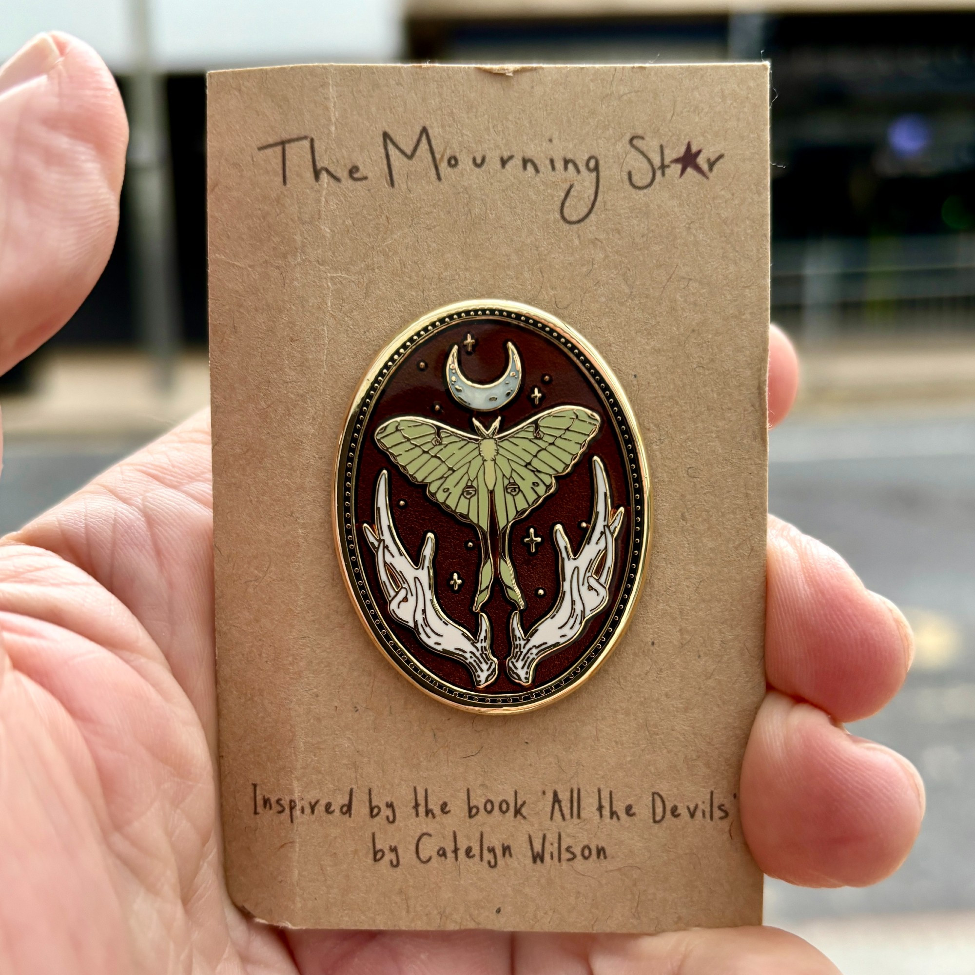 A pin on a cardboard backing, the pin has a crescent moon at the top with a lunar moth underneath then antlers at the bottom there are stars in the background of the glaze to go with the books 'All the Devils' by Catelyn Wilson