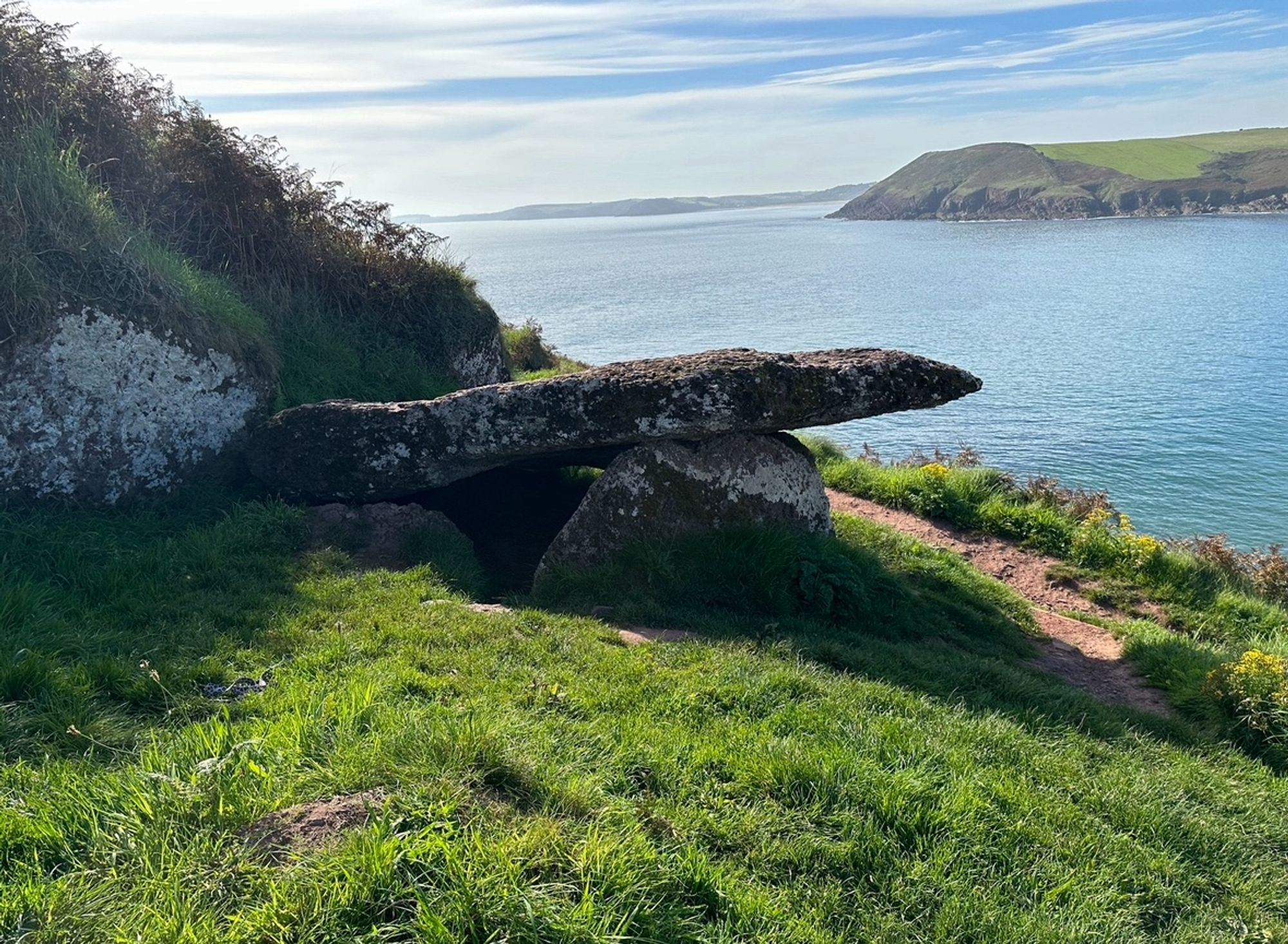 The chambered tomb viewed from the side with the open sea behind. The grass in the foreground is lush and green. The sea is textured by the gentle breeze. You can see ‘into’ the tomb from this angle through a gap in the upright stones supporting the capstone.