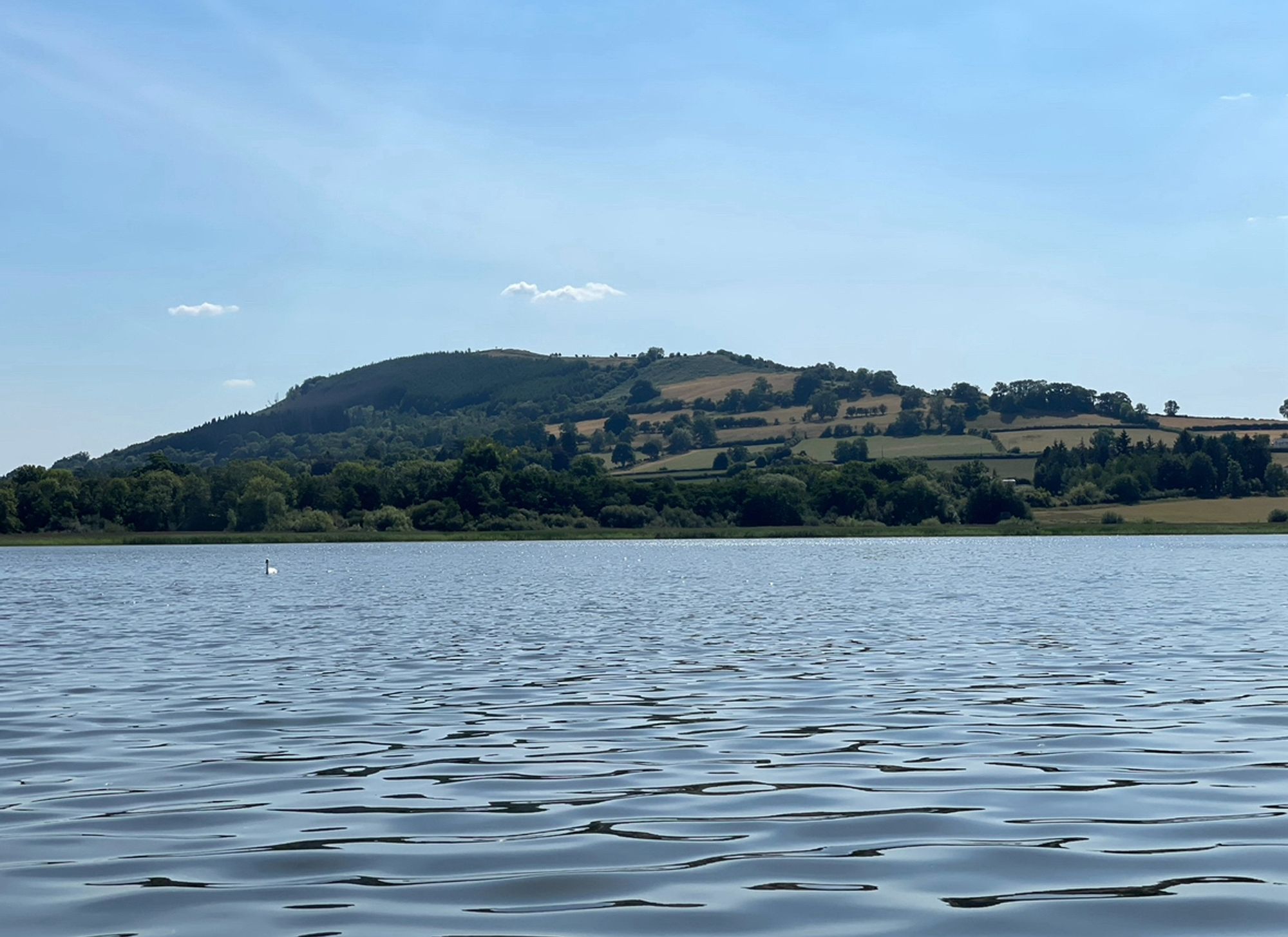 Zoomed in shot of the Hillfort emerging just beyond the lake. Some small white clouds hover near the top of the hill. New growth forest covers the Hillfort on one side. Cleared fields with hedgerow boundaries cover the other.