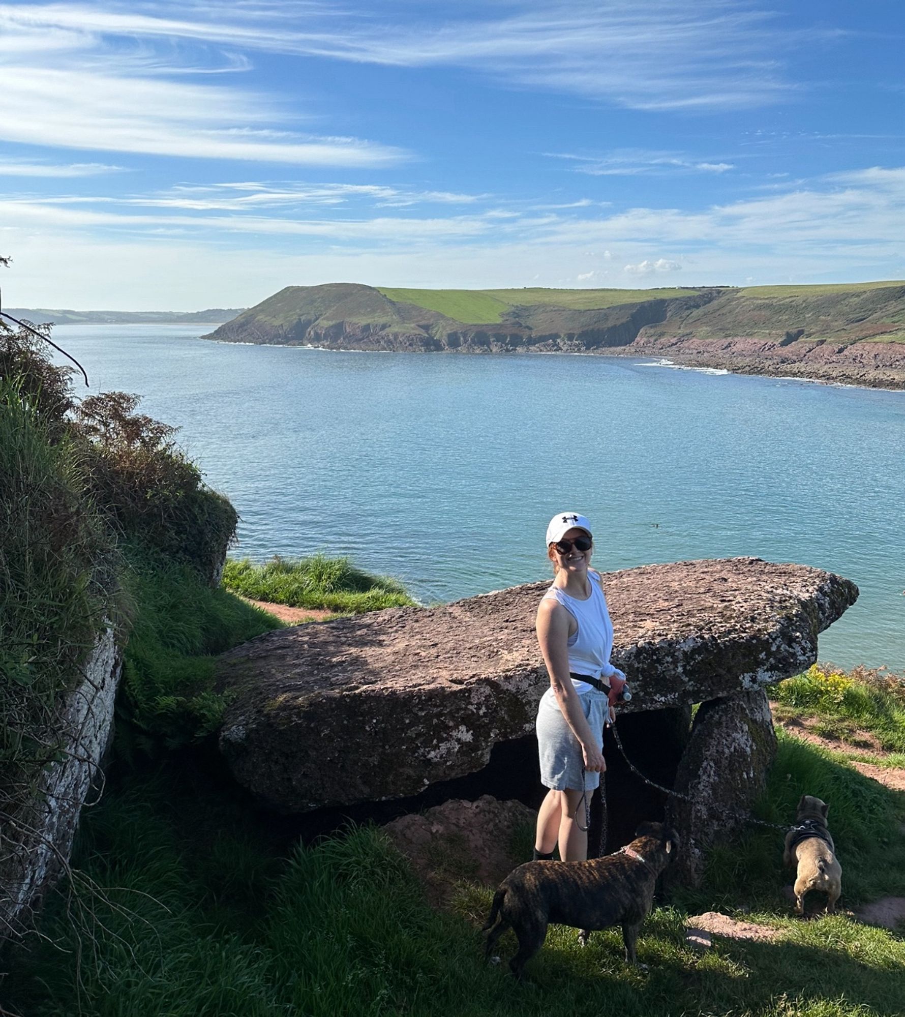 Me standing in front of the chambered tomb with my staffy and French bulldog on leashes. The capstone comes up to my chest and is angled slightly upwards towards the sea. It is partly underground. The sea behind is blue and calm. The blue sky has wispy white clouds.