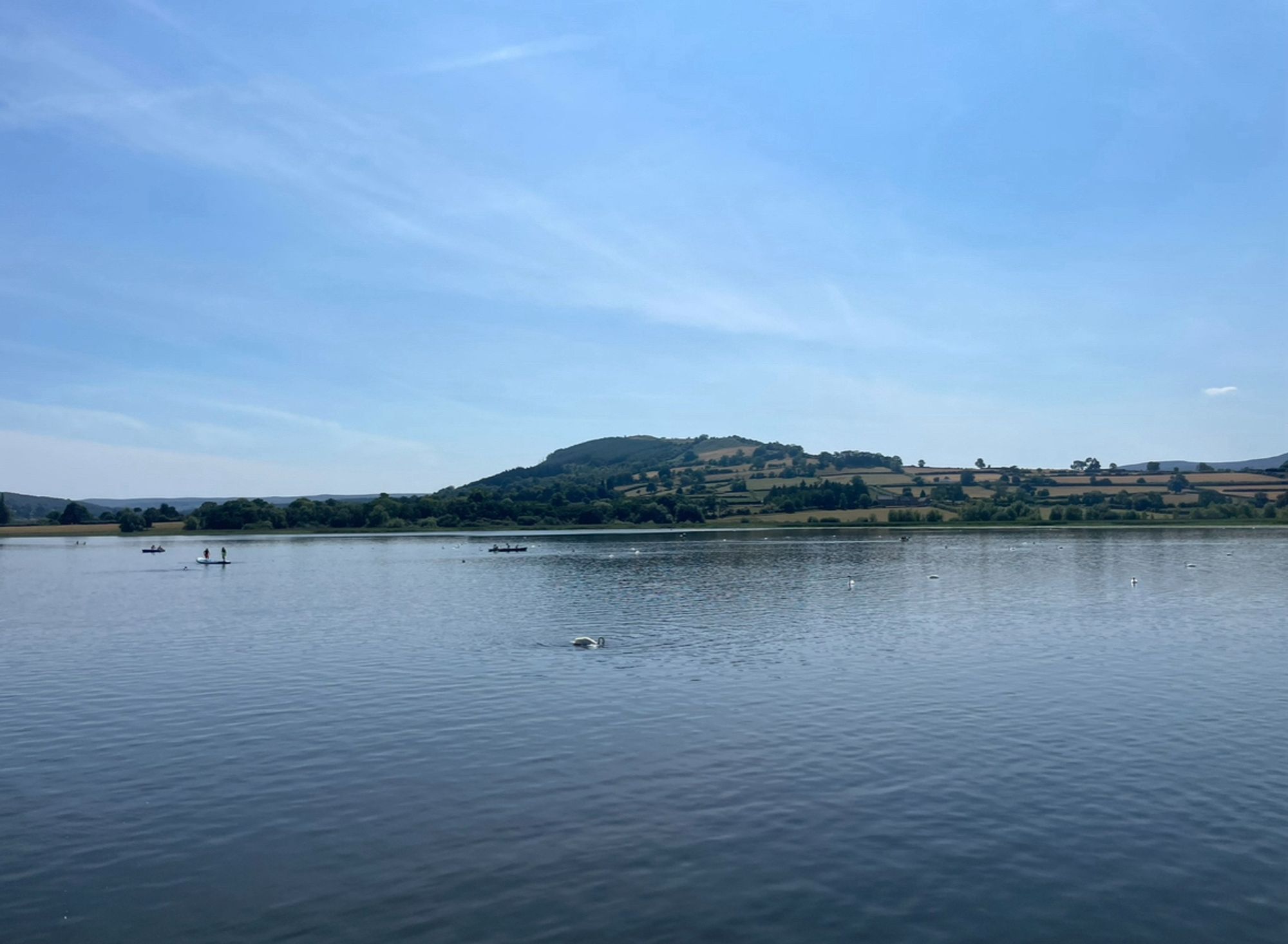 Photo of the calm lake waters and Hillfort in the background. Blue sky with wispy white clouds.