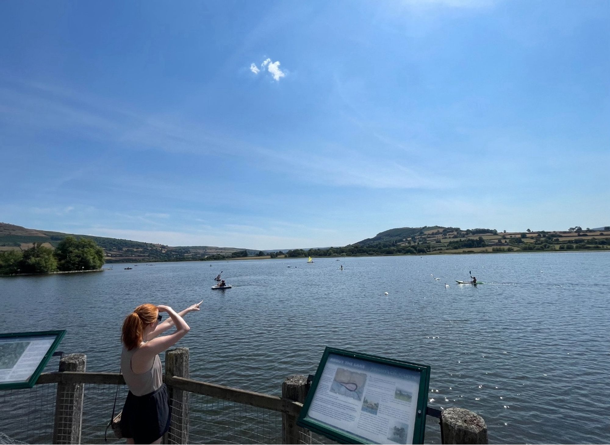 Me photographed from the side pointing across Llangors Lake to the Hillfort in the distance. It’s a sunny day. A couple kayakers paddle in the middle distance.