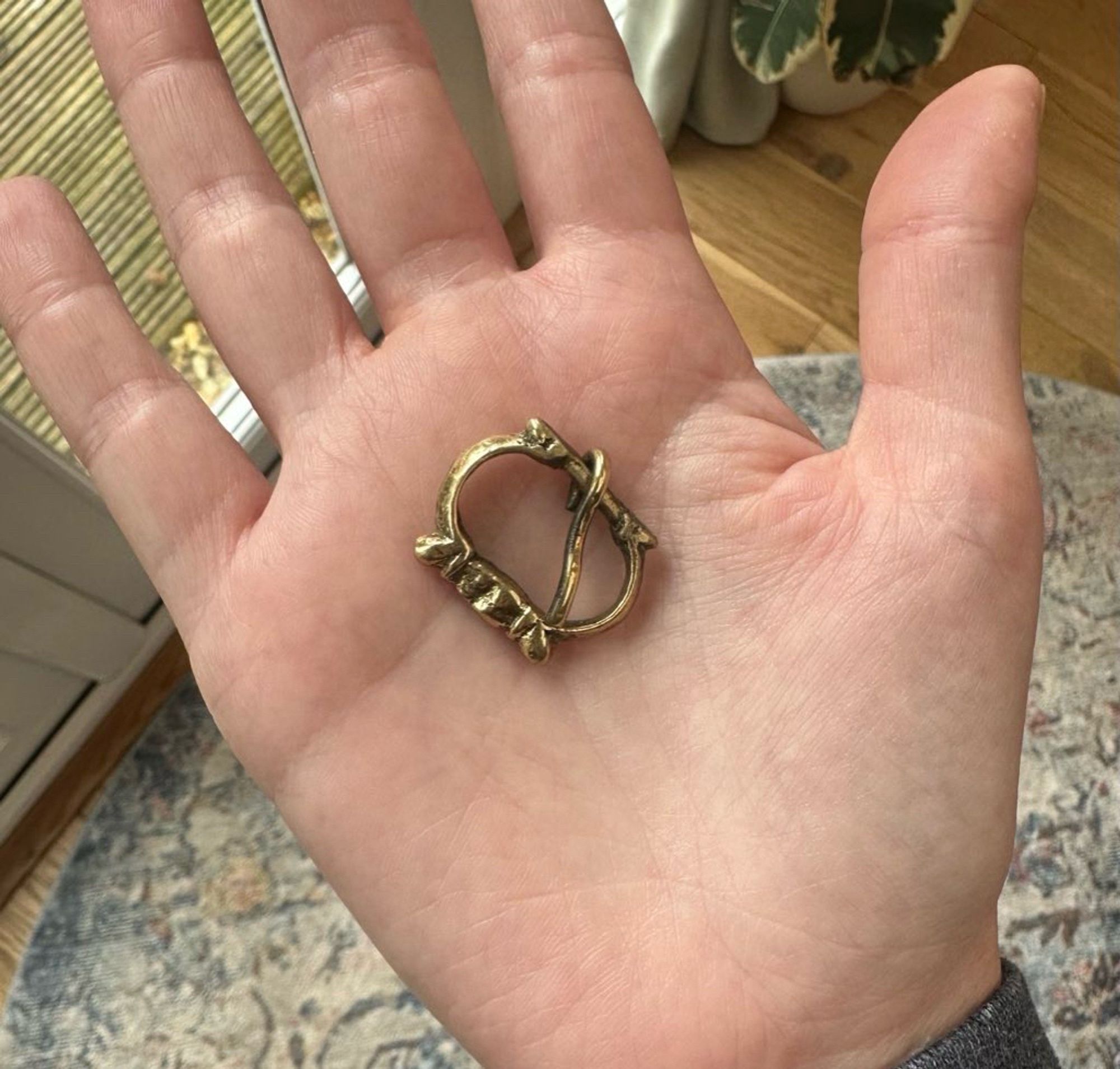 Photo of a replica brass buckle frame in the palm of my hand. It’s oval with a narrow offset bar and knobs flanking the pin rest.