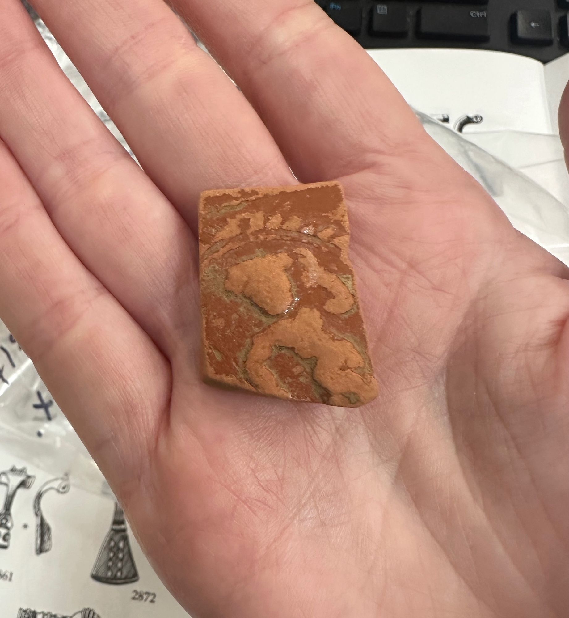 A sub-rectangular sheet of orange Samian ware in the palm of my hand. The sherd has a darker orange glaze. The sherd is decorated with a crude figure that is now unglazed from abrasion/erosion so it stands out against the dark orange background. The figure has one arm bent up at the elbow with hand near the head, the other arm bend down at the elbow like the lady in a red dress dancing emoji.