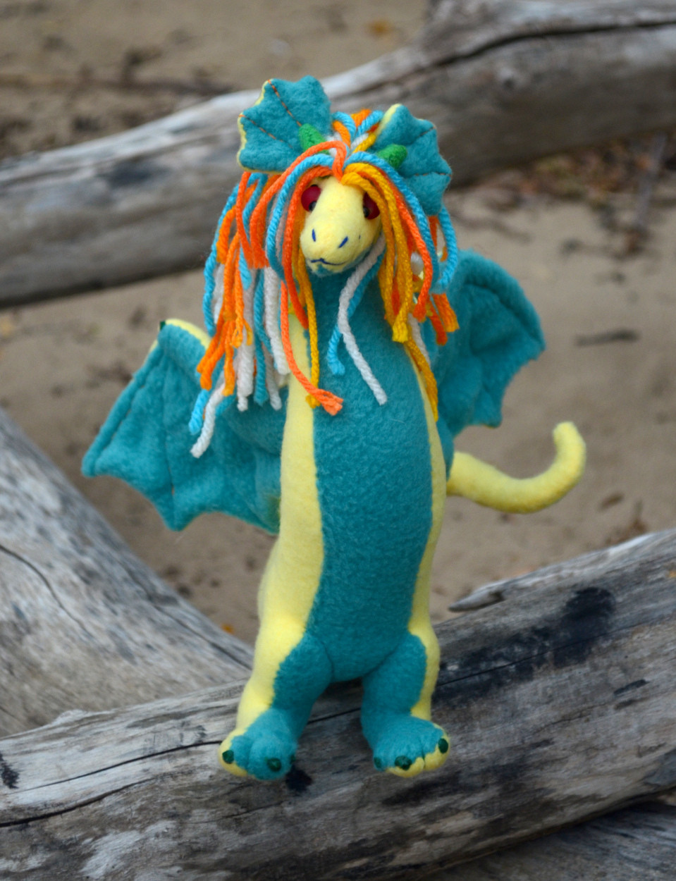 a 2 legged dragon plushie, light yellow with teal underbelly/details, an orange/teal/white mane, small green horns and claws, and red glass bead eyes