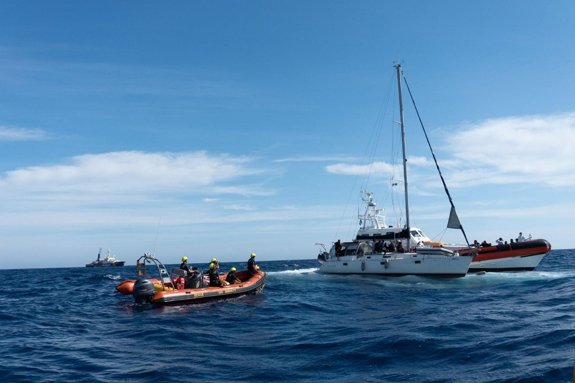 Ein Schnellboot der Humanity 1 vor einem Segelboot mit fliehenden Menschen an Bord auf hoher See. Dahinter ein Schnellboot der italienischen Küstenwache. Am Horizont die Humanity 1